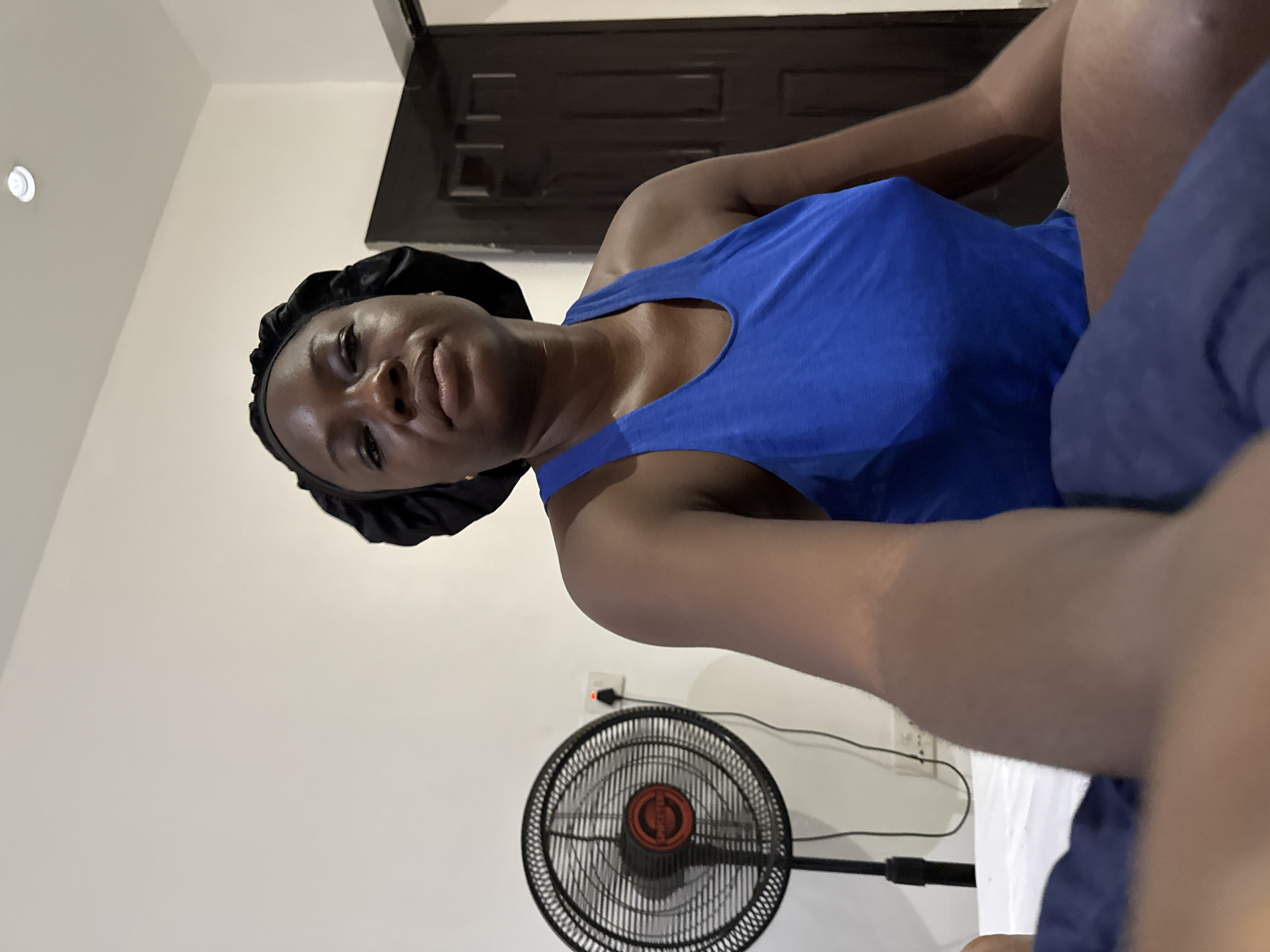 An adult female is captured in a selfie taken indoors, likely in Iddo Tudunwada, Nigeria. She has dark skin and looks directly at the camera with a neutral, relaxed expression. She wears a black satin bonnet and a blue sleeveless top. The image is taken from a low angle, suggesting the person is holding the camera herself, possibly while lying down or reclined, and the photo is rotated.
The background features a light-colored wall and ceiling. A white recessed light fixture is visible on the ceiling in the upper left. Behind the person's head and shoulder, there's a dark brown wooden door or piece of furniture. A black standing fan with an orange central hub is positioned in the lower part of the frame, plugged into a white wall outlet which displays a small red indicator light. The lighting in the room appears to be artificial. No text is visible in the image.