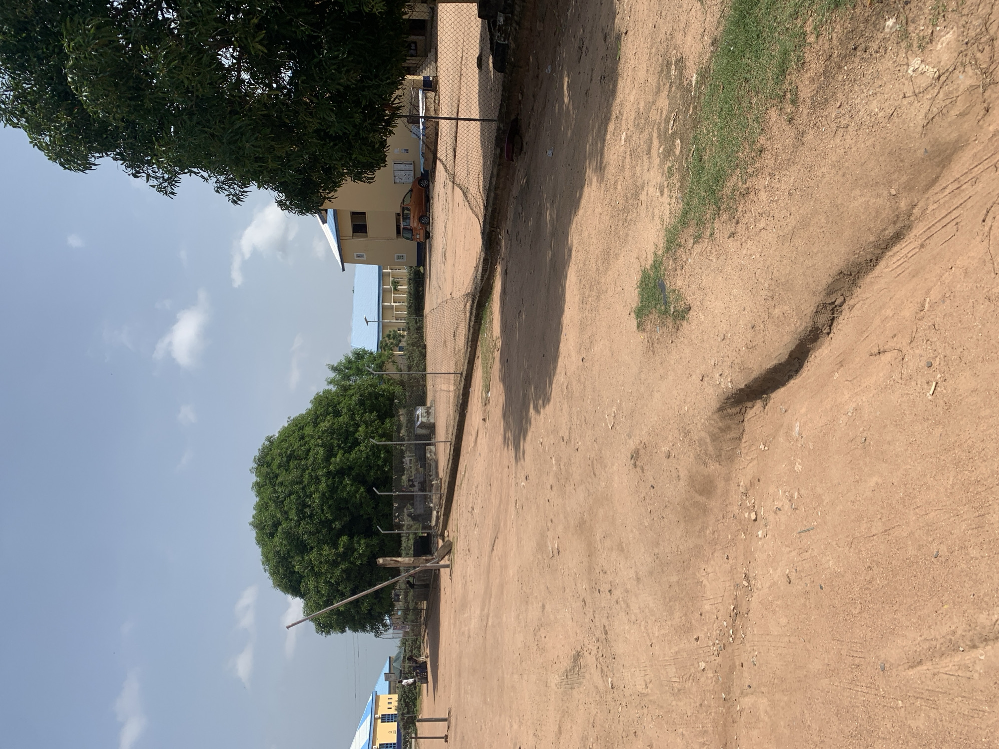The image depicts an outdoor scene on a dirt road in Jalingo, Nigeria. The foreground is dominated by the reddish-brown dirt road, which shows tire tracks and some erosion. To the left, a tall, thin wooden pole stands upright. In the mid-ground, a chain-link fence runs horizontally across the scene, separating the road from an area with some buildings and vegetation.
Behind the fence, there are several structures. A large, leafy green tree stands prominently in the left mid-ground. Further back, to the left, a building with a blue roof and yellow facade is visible, with a few people standing near it. To the right of this building, another structure with a yellow facade and a blue roof can be seen. Between these buildings, an orange car is parked.
The background features a clear blue sky with a few scattered white clouds. The overall environment appears to be somewhat arid, consistent with many parts of Nigeria. The lighting suggests it is daytime, likely under a bright sun given the shadows cast. The scene has a general appearance of a school or institutional compound, given the buildings and fence.