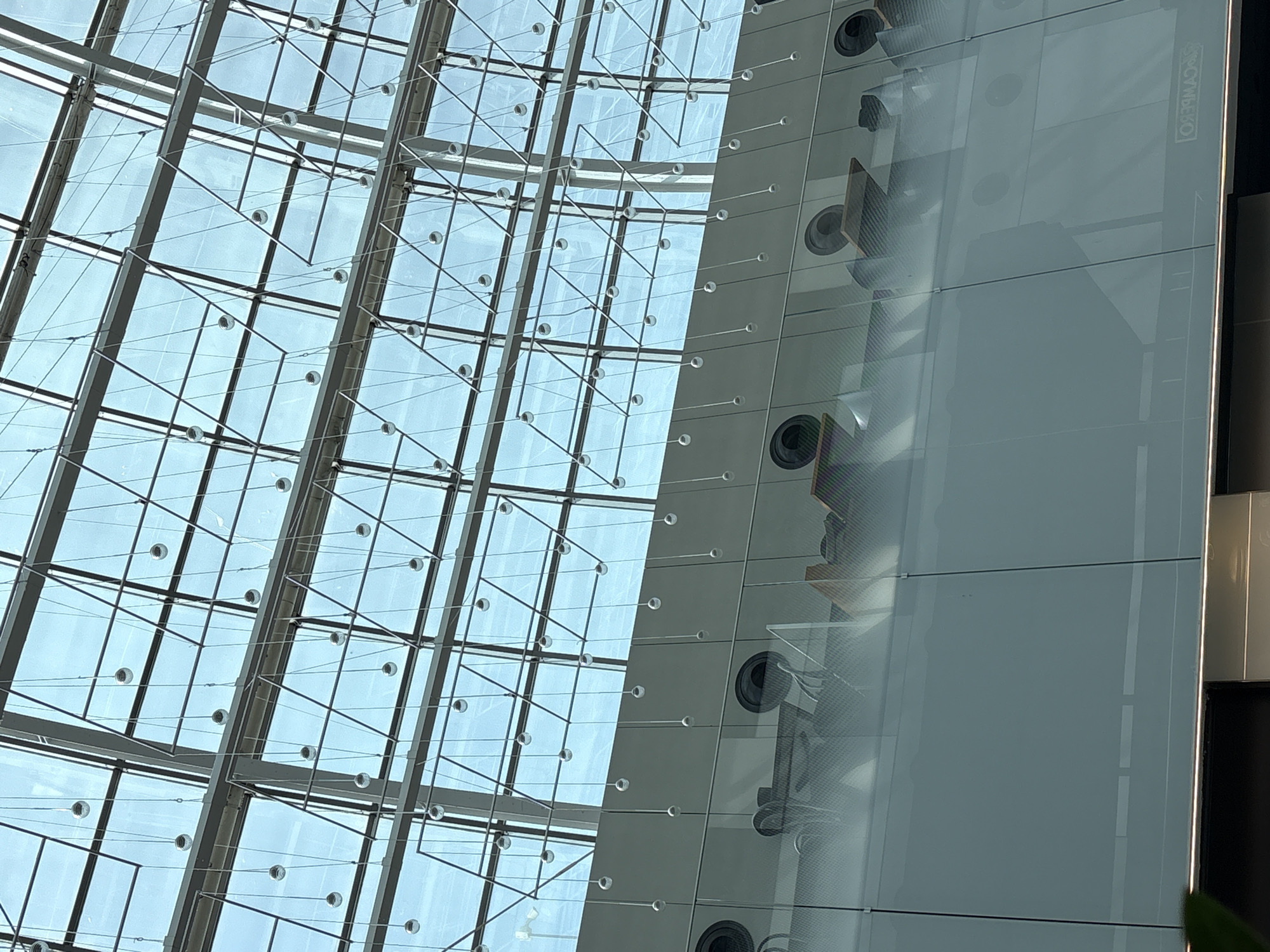 The image depicts the interior of a modern building, likely a large public space such as an airport terminal or convention center, given the expansive glass ceiling and architectural design. The ceiling is a complex grid of glass panes supported by a metallic framework, with thin wires extending downwards, each holding a small, spherical object, possibly decorative lights or some form of modern art installation.
Below the ceiling, a horizontal band of translucent material separates the upper and lower sections of the interior. This band features a series of circular openings, possibly ventilation grilles or decorative elements. Reflected in the glass of the lower section, we can see glimpses of a seating area with chairs and tables, suggesting that this area is designed for waiting or dining. The lighting is diffused, likely from the natural light filtering through the glass ceiling. The overall impression is one of spaciousness, contemporary design, and a clean, bright environment.