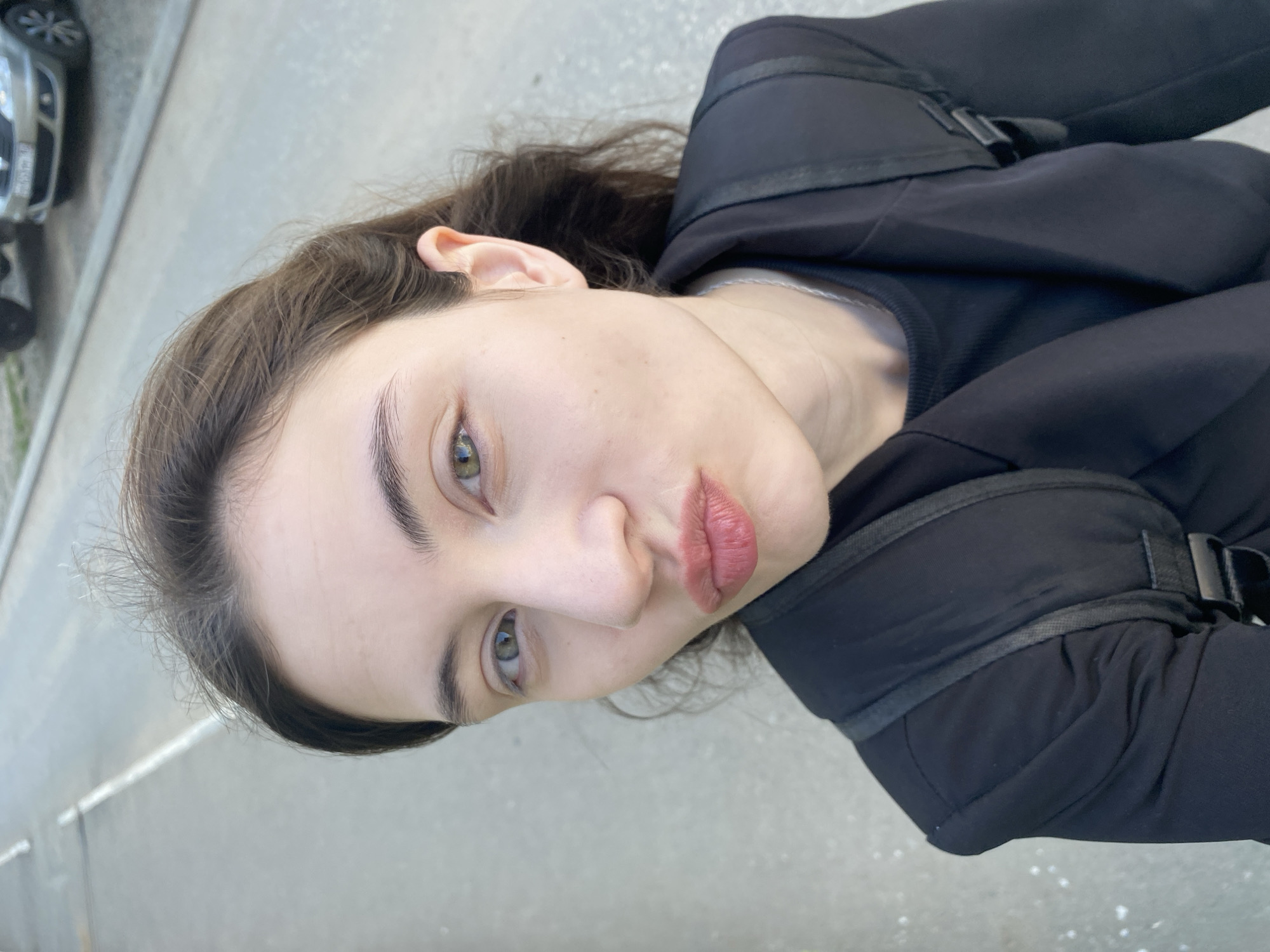 A young woman is the primary subject, wearing a black top and jacket with visible straps. She is facing the camera with a slight tilt of her head. Her eyes are light, and her lips are a muted red. The background consists of a grey concrete surface with a road marking, suggesting an urban setting. In the upper left corner, a portion of a parked silver car is visible. Given the context of Votkinsk, Russia, the scene likely takes place on a street or sidewalk within the city limits, though specific landmarks are not discernible in the image. There are no apparent actions or interactions in progress. The environment appears to be outdoors, with natural lighting.