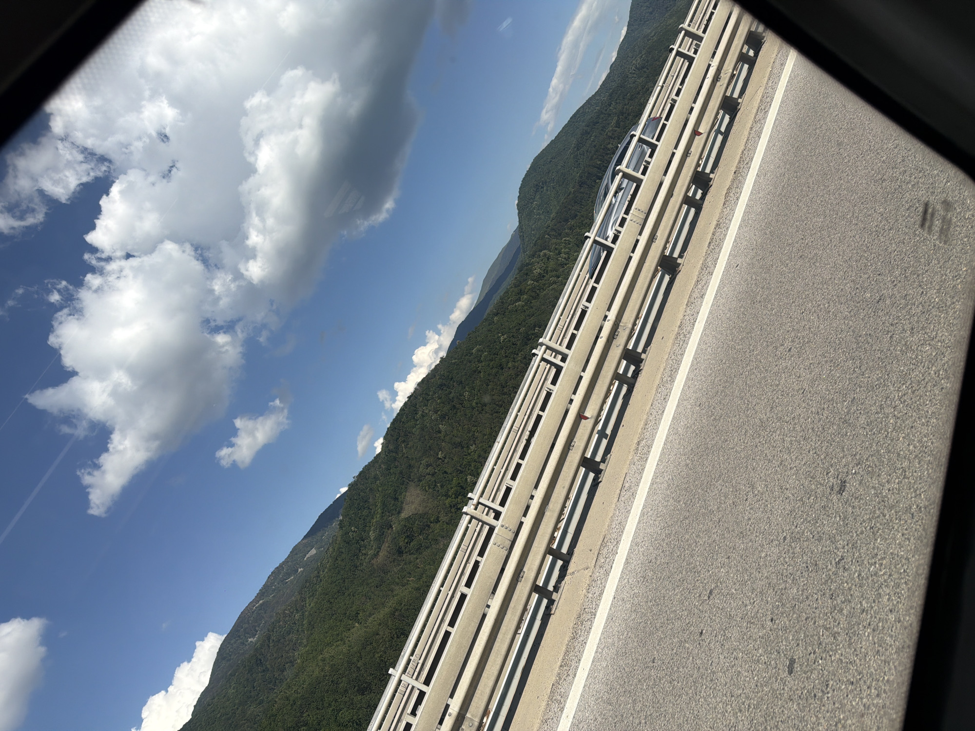 This media file captures a scenic view from a car driving on a road, likely a bridge or elevated highway, in Vrachesh, Bulgaria. The perspective is tilted, suggesting the photo was taken from a passenger's window.
The main subjects are the road itself, a metal guardrail, a densely forested mountainside, and a bright blue sky filled with fluffy white clouds. A light-colored car is visible in the distance, driving on the same road, partially obscured by the guardrail.
The scene depicts a daytime drive through a mountainous landscape. The sun is shining, casting shadows from the guardrail onto the road, indicating a clear and pleasant day. The lush green of the trees suggests it is likely spring, summer, or early autumn. The mountains are tall and imposing, with the road winding along their side.
Notable details include the modern construction of the guardrail, composed of parallel metal bars and concrete supports. The asphalt road has a clear white line marking the lane. The clouds are dynamic, with some appearing wispy and others more substantial, hinting at a light breeze. There are no visible signs of text in the image. The overall mood is one of peaceful travel and appreciation for natural beauty.