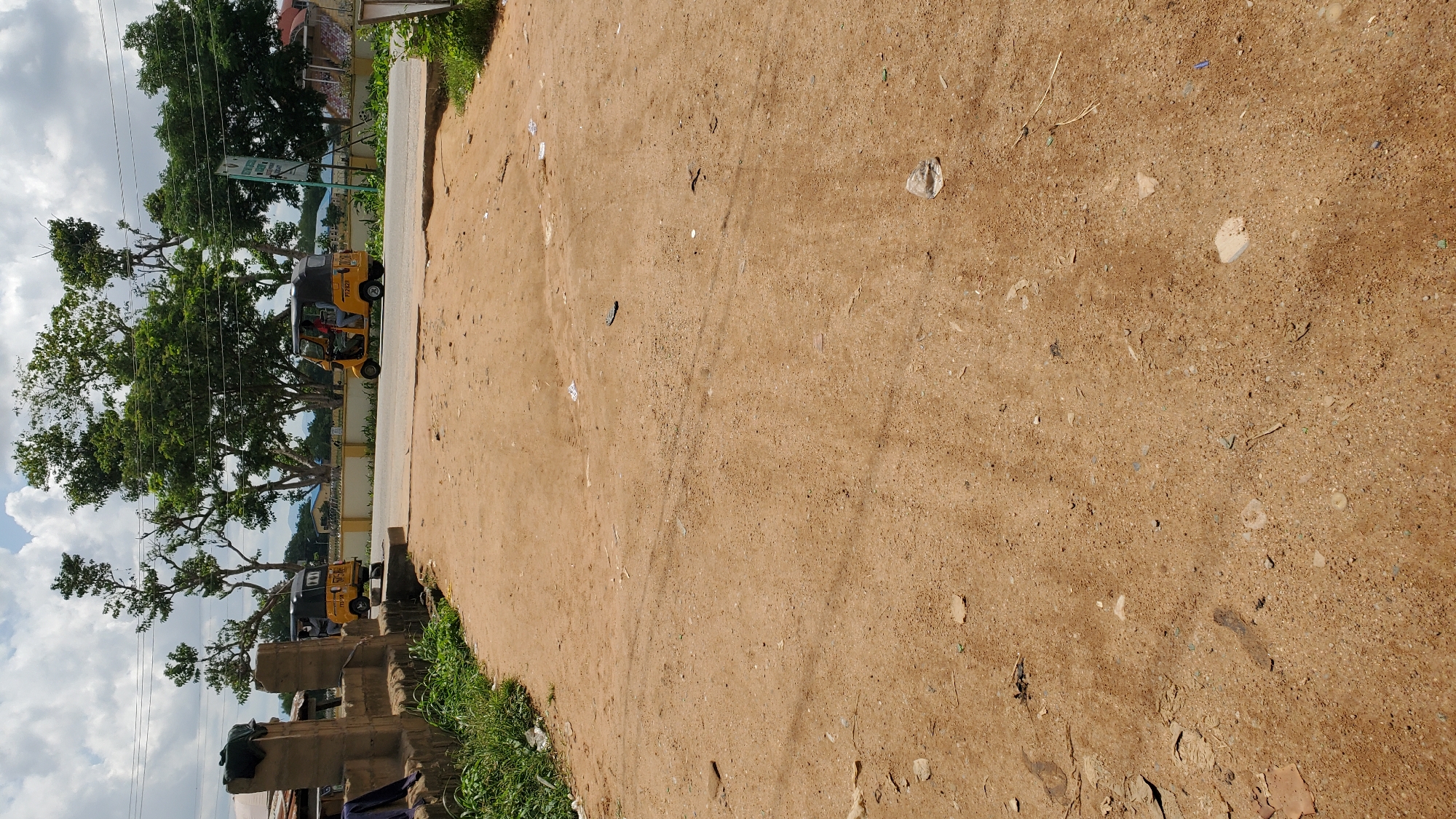 A medium shot captures an outdoor scene in Jalingo, Nigeria, on a cloudy day. The foreground is dominated by a dirt path with scattered debris, including plastic bags and small rocks. In the midground, a paved road runs horizontally, with two yellow and black tuk-tuks driving on it. The tuk-tuk on the left is seen from the rear, while the one on the right is viewed from the side, showing passengers. Beyond the road, a yellow and white striped wall encloses a grassy area and trees with lush green foliage. In the background, buildings and more trees are visible, with mountains in the distance. The overall impression is one of a typical urban or semi-urban environment in Nigeria, with a mix of natural and man-made elements.