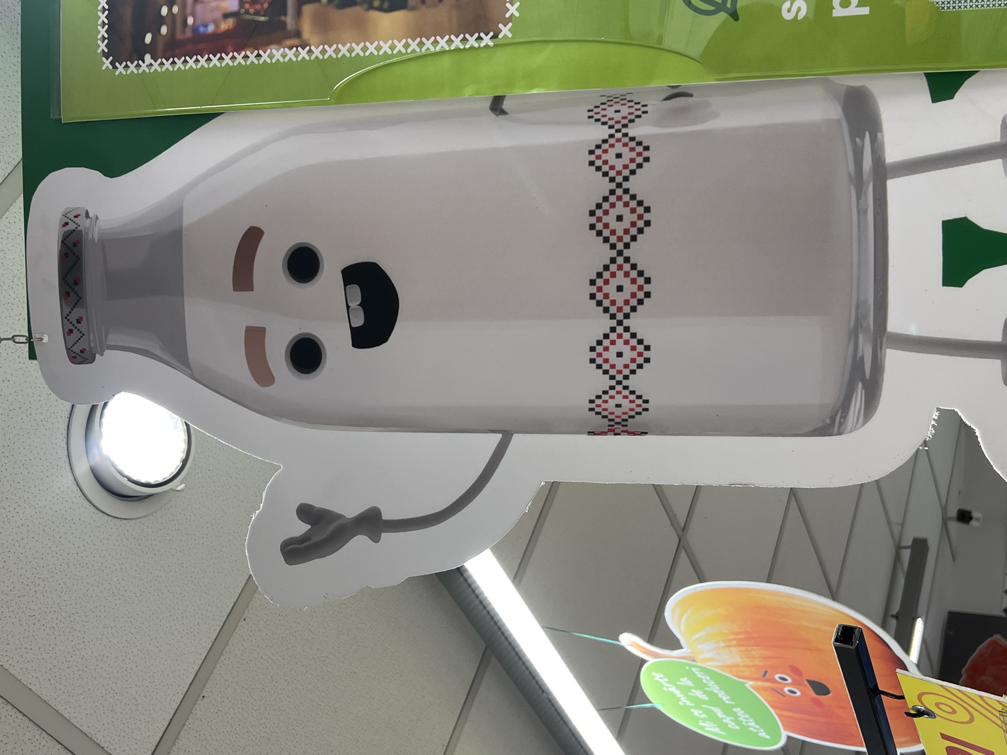 The image shows a close-up view of a large, cartoonish cutout of a milk bottle with a face, hanging from a ceiling in what appears to be a retail store. The milk bottle cutout is white with a grey cap that has a decorative pattern in red, black, and white. It has two black circular eyes, a wide black mouth with two white teeth, and two brown eyebrows. A grey, stylized arm with a hand is visible to the left of the bottle, reaching upwards.
To the left of the milk bottle cutout, a large, orange cutout shaped like a pumpkin or squash is visible. It has a smiling face with two eyes and a mouth, and a green speech bubble attached to it that contains text in a foreign language. Below the pumpkin cutout, a portion of a yellow sign with text is visible.
The environment is a store interior with a suspended ceiling and bright fluorescent lighting. A green display board with white decorative stitching is visible to the right of the milk bottle. The overall impression is a whimsical display, likely for promotional purposes. The context provided by the user suggests this scene is located in Chișinău, Moldova.