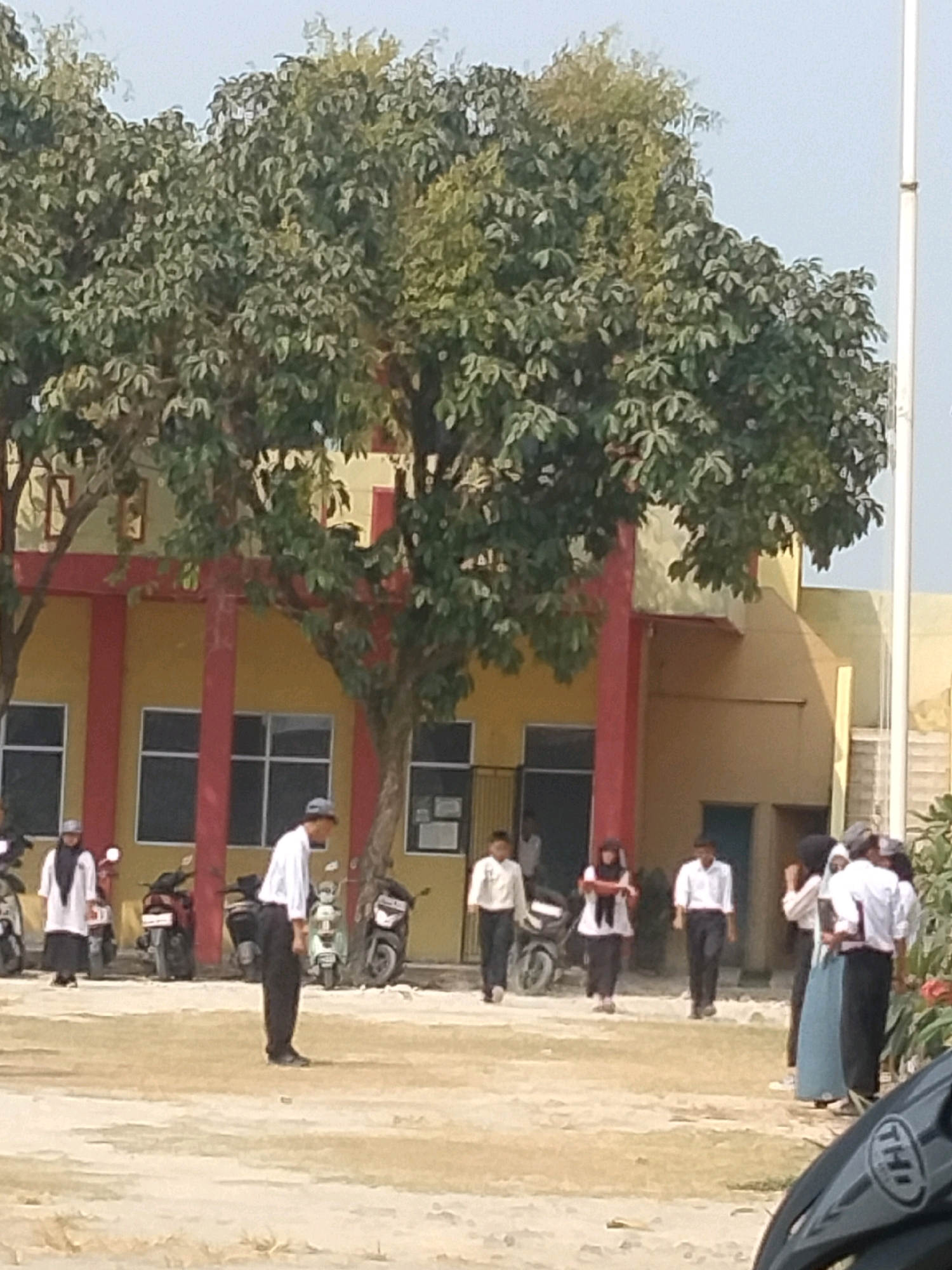 The image depicts an outdoor scene in Suka Sari, Indonesia, likely in front of a school building. A yellow building with red trim and pillars forms the backdrop. Several individuals are visible, including students and possibly staff. On the left, a person wearing a white hijab and black attire stands near a motorcycle. In the center, a man in a white shirt and black pants is walking, and another person, also in a white shirt and black pants, walks towards the right. To the right, a group of individuals in similar school uniforms are gathered. Motorcycles are parked in the background, suggesting a mode of transportation common in the area. Large trees with green foliage partially obscure the building and the sky. The ground appears to be unpaved. The overall impression is a typical day at an educational institution in Indonesia.