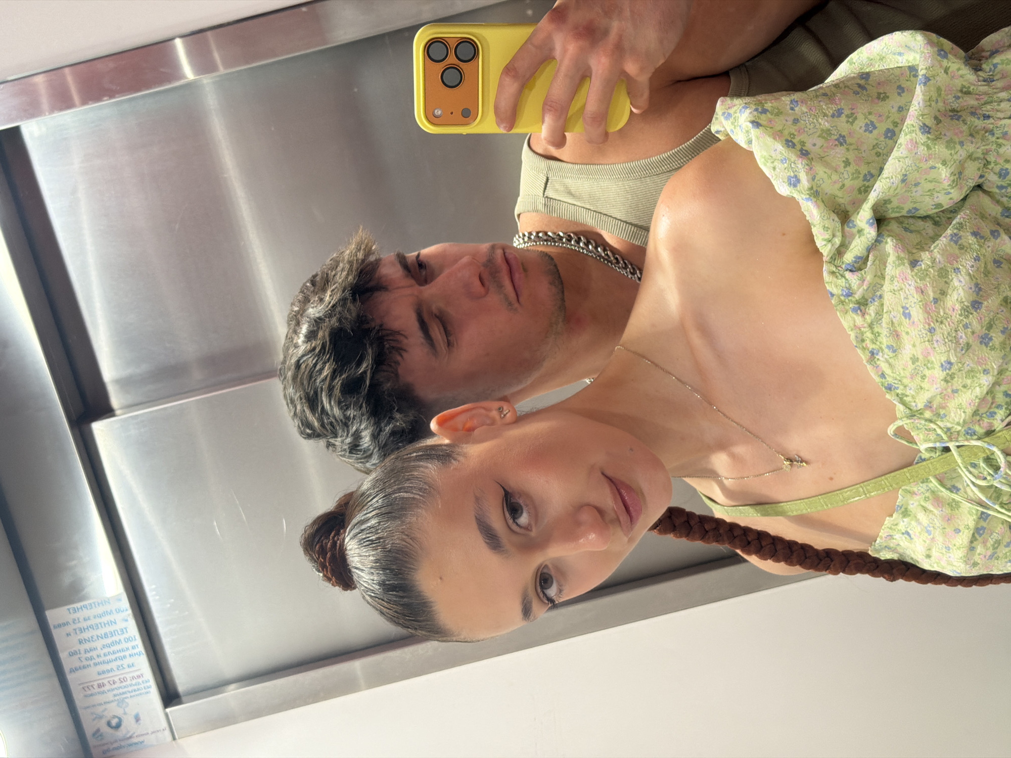 A young couple is taking a selfie in an elevator. The woman, in the foreground, has her hair pulled up in a bun with a long braid down her back. She is wearing a light green floral off-the-shoulder top and a gold necklace. The man, behind her, is holding up a yellow smartphone to take the picture. He is wearing a ribbed tank top and a thick silver chain necklace. The elevator has stainless steel doors and walls. A label with Cyrillic text is visible on the elevator wall, which is consistent with Sofia, Bulgaria.