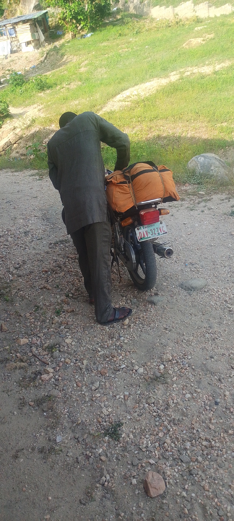A man wearing a dark suit and sandals is standing beside a motorcycle on a gravelly road. The motorcycle has an orange duffel bag strapped to the back and a Nigerian license plate reading "DAY-327VC." The man is leaning over the motorcycle, possibly adjusting something.
In the background, there is a grassy hill and a small, makeshift structure. This scene is likely taking place on the outskirts of the city of Lokoja, Nigeria.