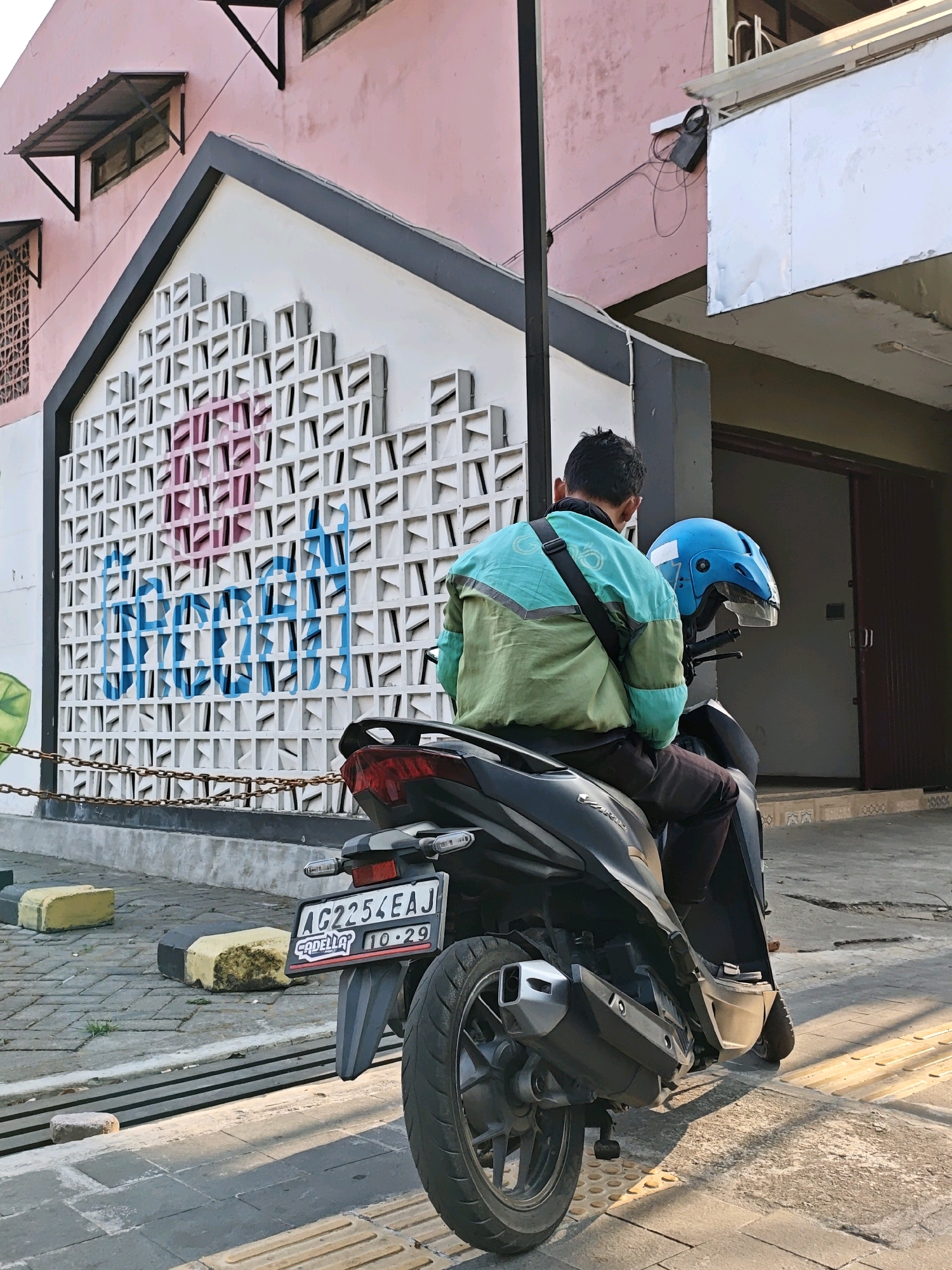 A person wearing a green and blue jacket is sitting on a black motorcycle, facing away from the camera. The motorcycle has a blue helmet on its handlebars and a license plate that reads "AG 2254 EAJ". Behind the motorcycle, there is a white wall with a patterned design and some blue and pink graffiti. The building has a pink facade and dark gray trim. The scene appears to be on a street with a brick-paved sidewalk. The overall lighting suggests it is daytime. The location is in the city of Kediri City, Indonesia, as indicated by the license plate format.