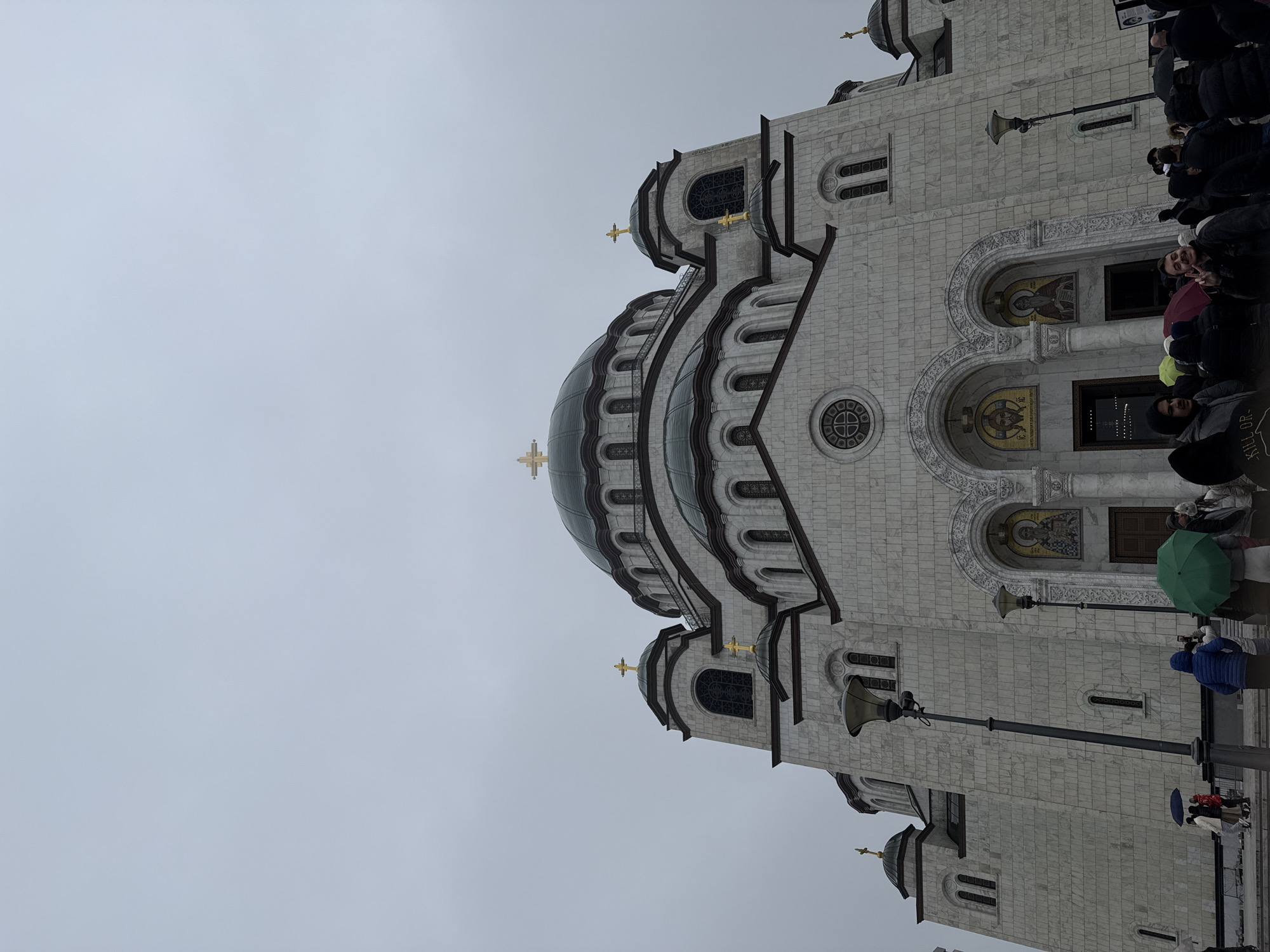 The image shows the Church of Saint Sava in Belgrade, Serbia. The church is a large, ornate building with a central dome and several smaller domes, all topped with gold crosses. The exterior is made of white marble. The sky is overcast. There are people near the church and an umbrella in the foreground. A street lamp is also in the foreground.