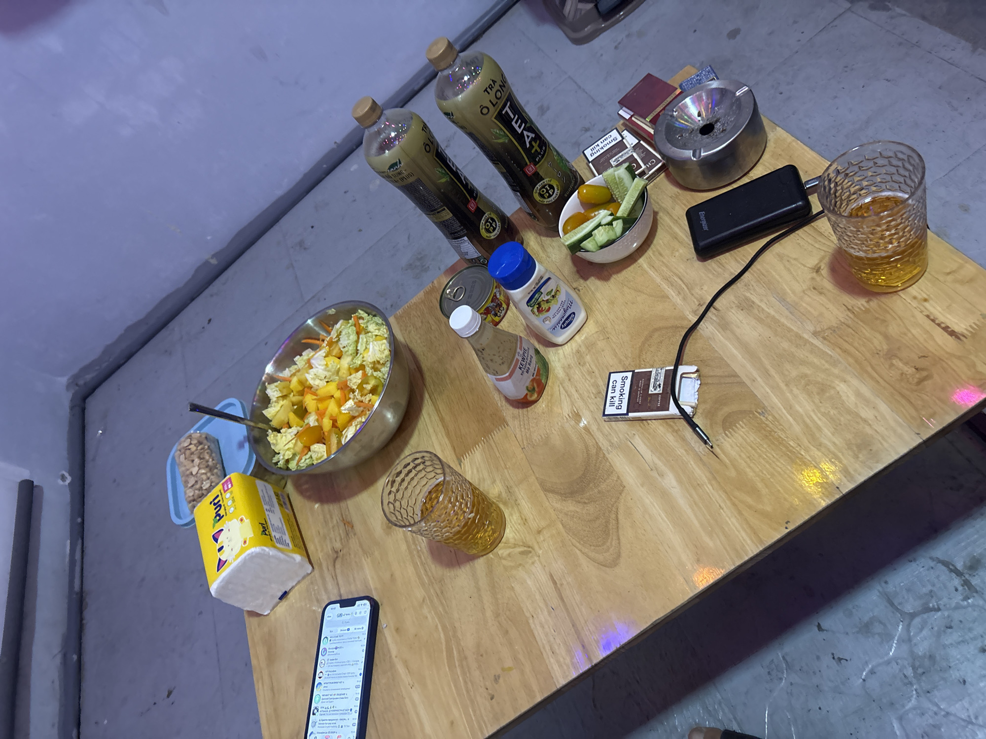 The image depicts a table in a dimly lit room, possibly in a residential setting. The location is Nha Trang Ward, Vietnam, based on the presence of Vietnamese-labeled products. The table is made of light-colored wood and holds a variety of items.
Food items include a large salad in a metal bowl, a small bowl with cucumbers and tomatoes, a small tray of peanuts, and a box of "Puri" brand tissues. There are two bottles of "TEA+" Oolong tea, a can of tuna, and bottles of Kewpie and mayonnaise.
Beverages consist of two glasses, each filled with a yellow liquid, likely beer or a similar drink. A black power bank is connected to a device by a black cable.
Other objects include a silver ashtray, a pack of cigarettes labeled "Smoking can kill", a mobile phone displaying a messaging app, and several small books. The floor around the table is tiled in grey. No people are clearly visible in the image.