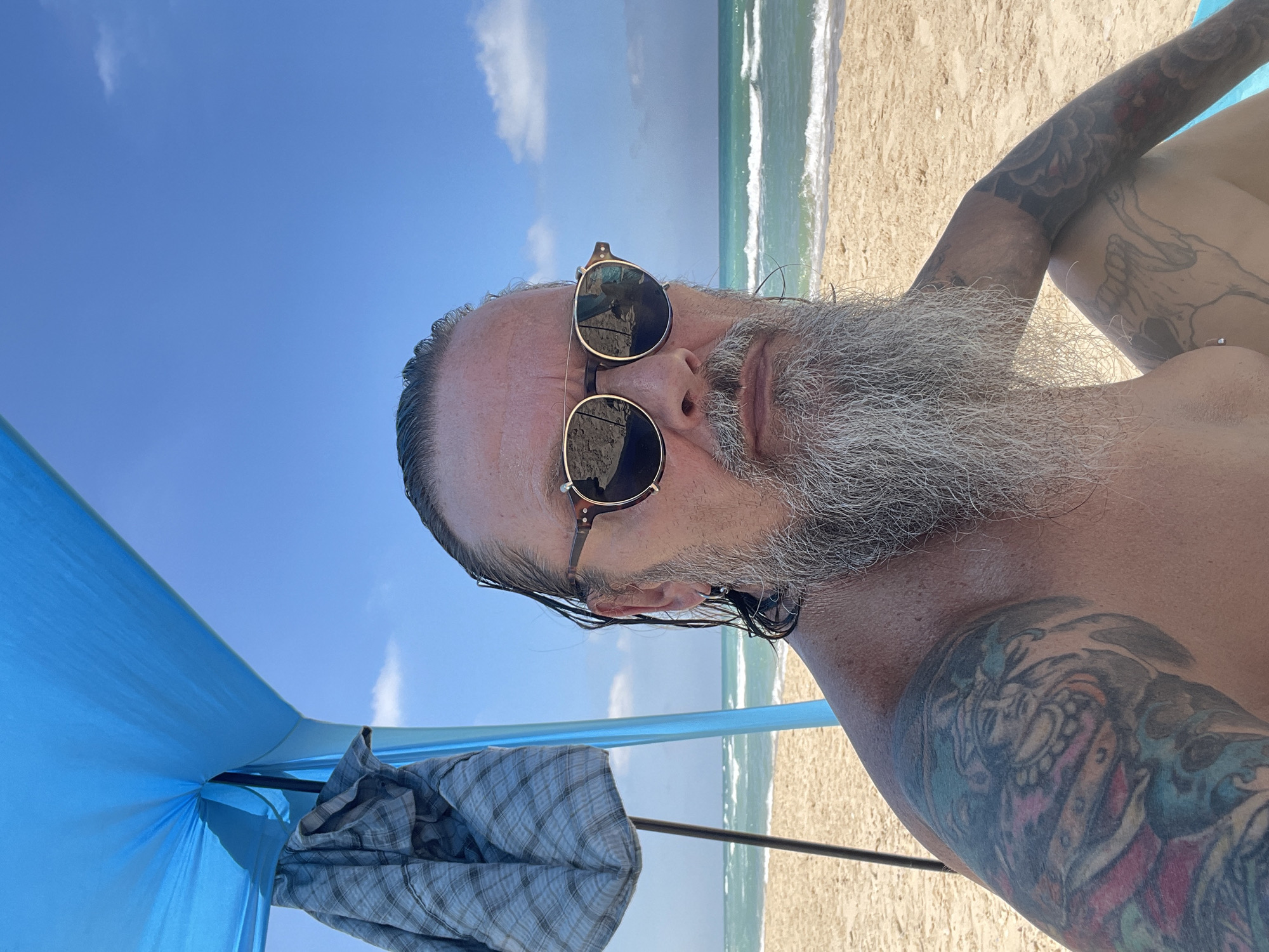 A man with long gray hair pulled back, a long gray beard, and sunglasses is seated on a sandy beach in Ga'ash, Israel, under a blue beach canopy. He is shirtless, revealing multiple colorful tattoos on his arms and a sketch-like tattoo on his leg. The ocean with moderate waves is visible in the background, under a clear blue sky with scattered clouds. A grey and blue plaid shirt is draped over a pole of the canopy. The man is looking directly at the camera.