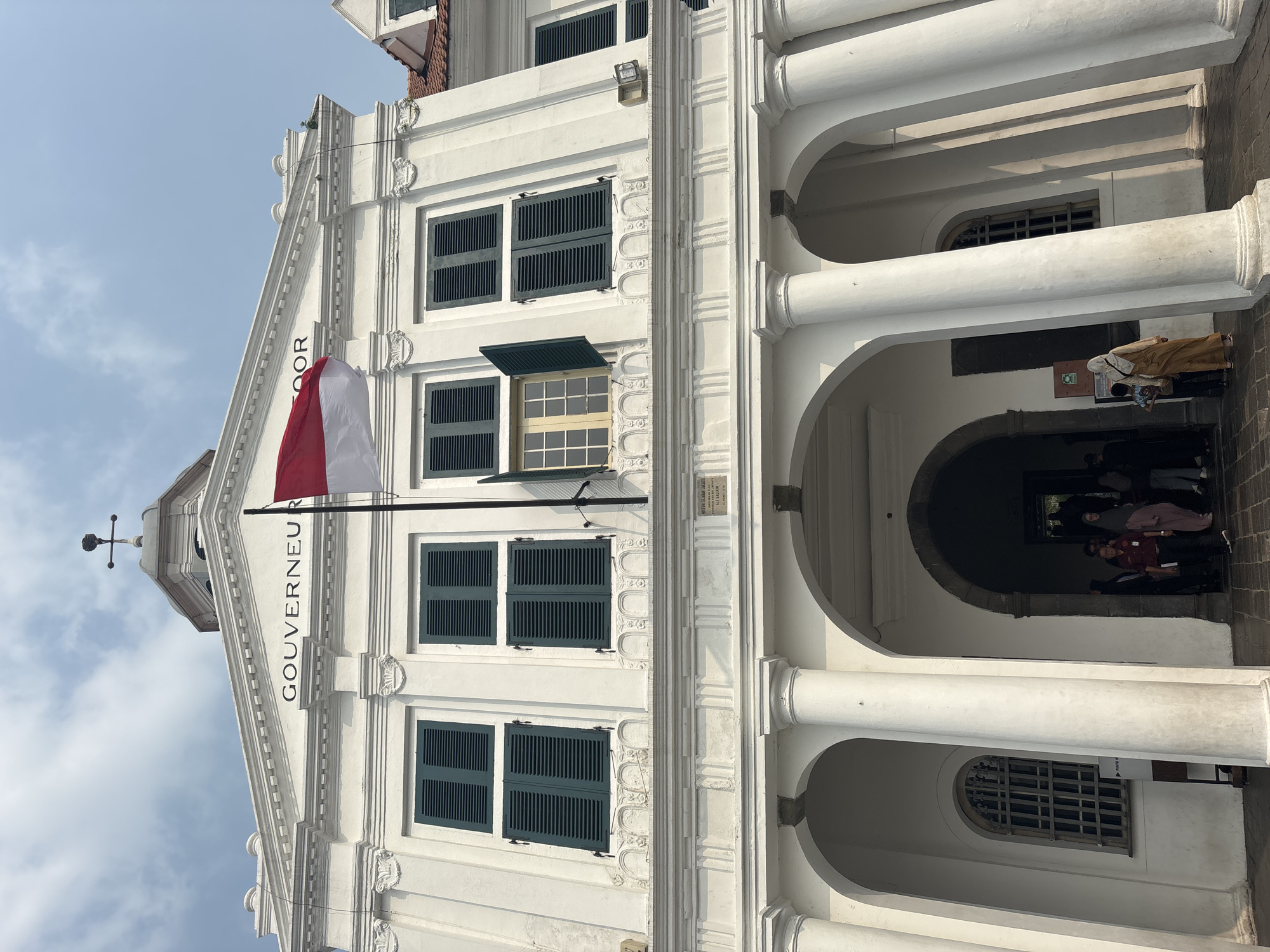 This is a vertical, eye-level, outdoor photograph of the Fatahillah Square building in West Jakarta, Indonesia, on a bright, cloudy day. The building is white with classical architectural features, including columns and arched doorways. A red and white Indonesian flag flies from a flagpole on the roof. Above the central entrance, the word "GOUVERNEUR" is inscribed on the facade. Several people are visible entering the building through an arched doorway. The ground is paved with dark cobblestones. The building's facade is symmetrical, with multiple windows on the upper floors, some of which have dark green shutters.