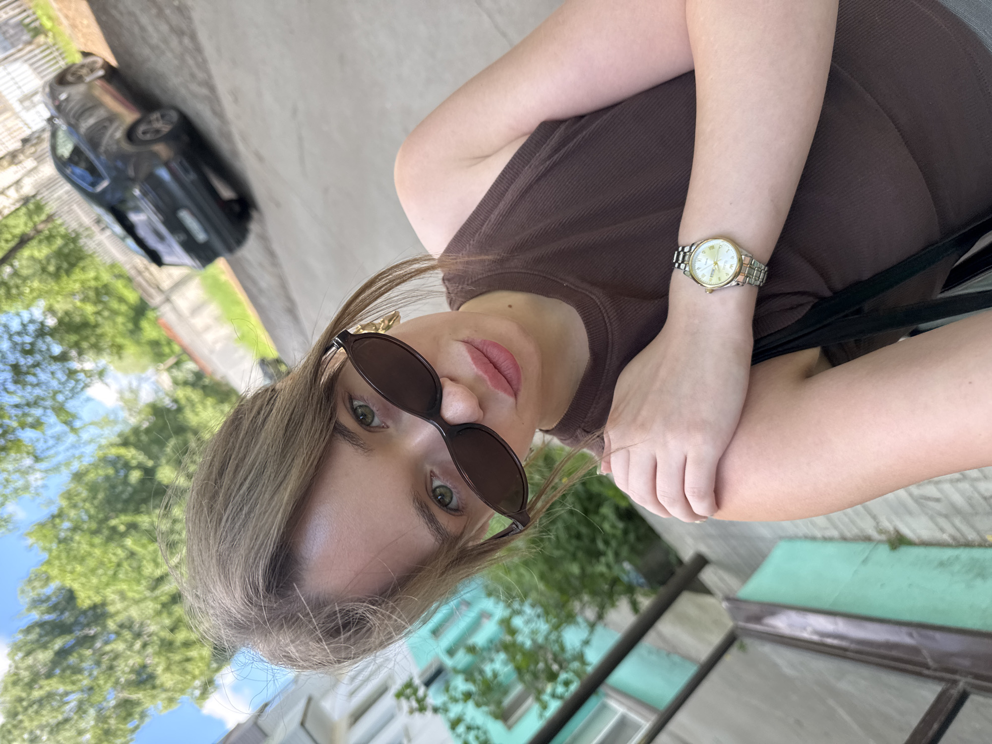 A young woman with light brown hair pulled back, wearing large dark sunglasses, a brown tank top, and a silver watch on her left wrist, poses for a selfie outdoors. She is looking directly at the camera with a slight pout. Her right arm is bent, with her hand resting on her left shoulder. In the background, a dark-colored SUV is parked on a paved surface, and a light-colored fence runs along the right side of the frame. To the left, a building with turquoise accents and green foliage is visible. The sky above is blue with scattered clouds and leafy trees. The scene appears to be in a residential area in Chaikovsky, Russia, with the architecture and vehicle suggesting a contemporary urban setting.