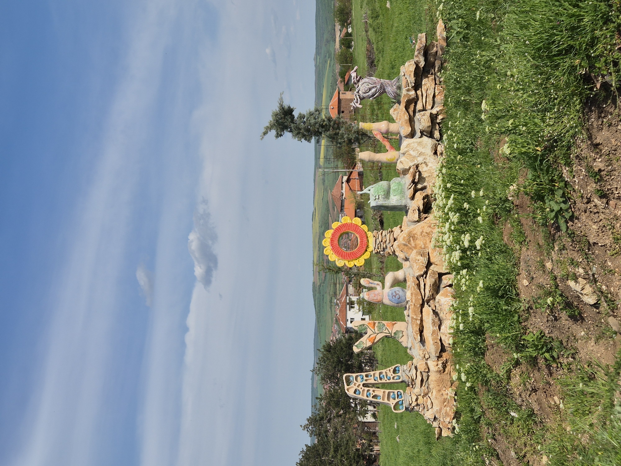 The image captures a scene in Lukovit, Bulgaria, showing an outdoor art installation made of stone and concrete figures, including human-like figures and geometric shapes, located on a hillside. Behind the artwork, the landscape includes green fields, scattered rural houses with red-tiled roofs, and distant rolling hills. The sky is blue with some white clouds. The immediate foreground consists of grassy vegetation and a rocky, dirt embankment.