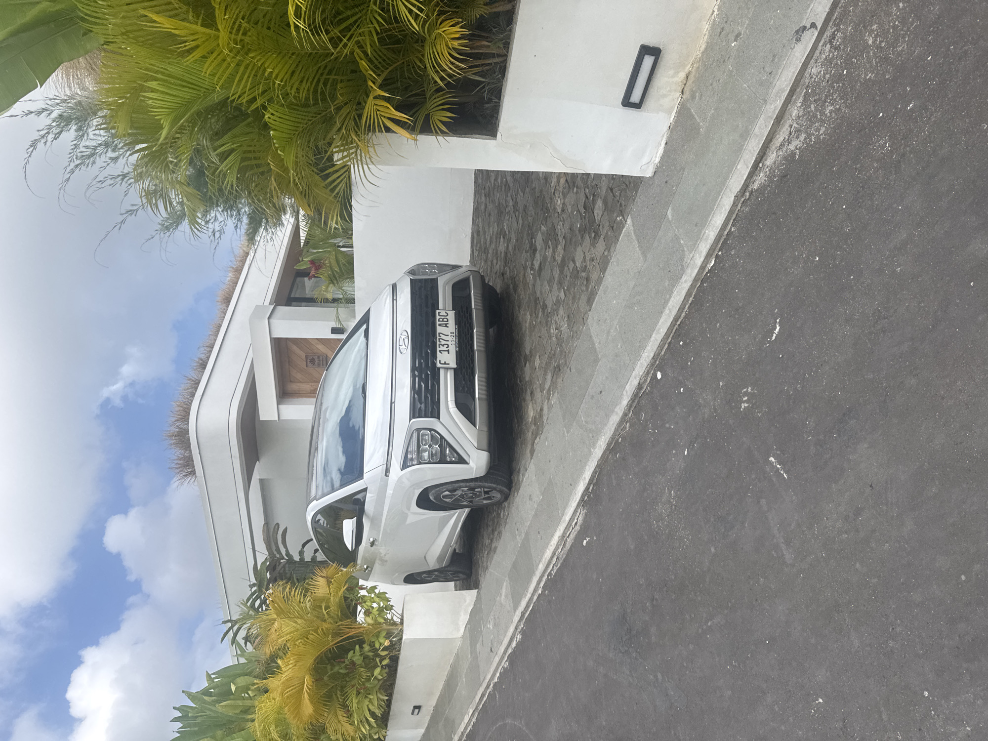 A white Hyundai Creta is parked on a patterned stone driveway in front of a modern white building with a green roof. Palm trees with lush green and yellow fronds surround the building. The sky above is blue with scattered white clouds, suggesting a bright, sunny day. A black rectangular light fixture is mounted on the white wall of the building. The car's license plate reads "F 1377 ABC". The scene appears to be in a tropical or sub-tropical location, consistent with Kuta Selatan, Indonesia. The overall impression is one of a well-maintained, modern property in a scenic, sunny environment.