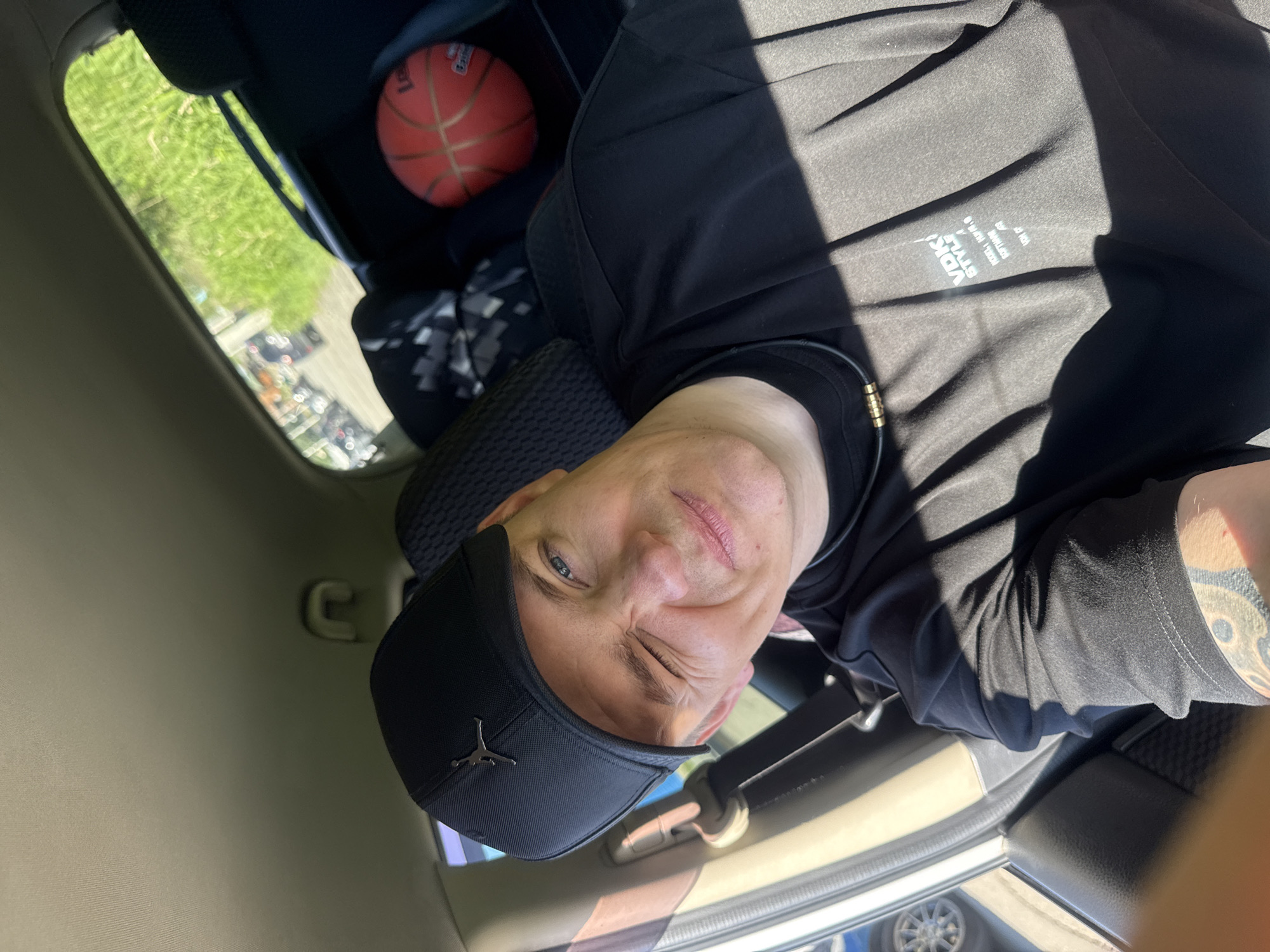 A man wearing a black baseball cap and a black t-shirt is sitting in the passenger seat of a car, taking a selfie. He is winking at the camera. A basketball is visible on the back seat of the car. The scene appears to be outdoors, possibly in a city, as cars are visible through the window. The lighting suggests it is daytime. The man has a tattoo on his arm. The image does not provide any information to suggest the location is Vladivostok, Russia.