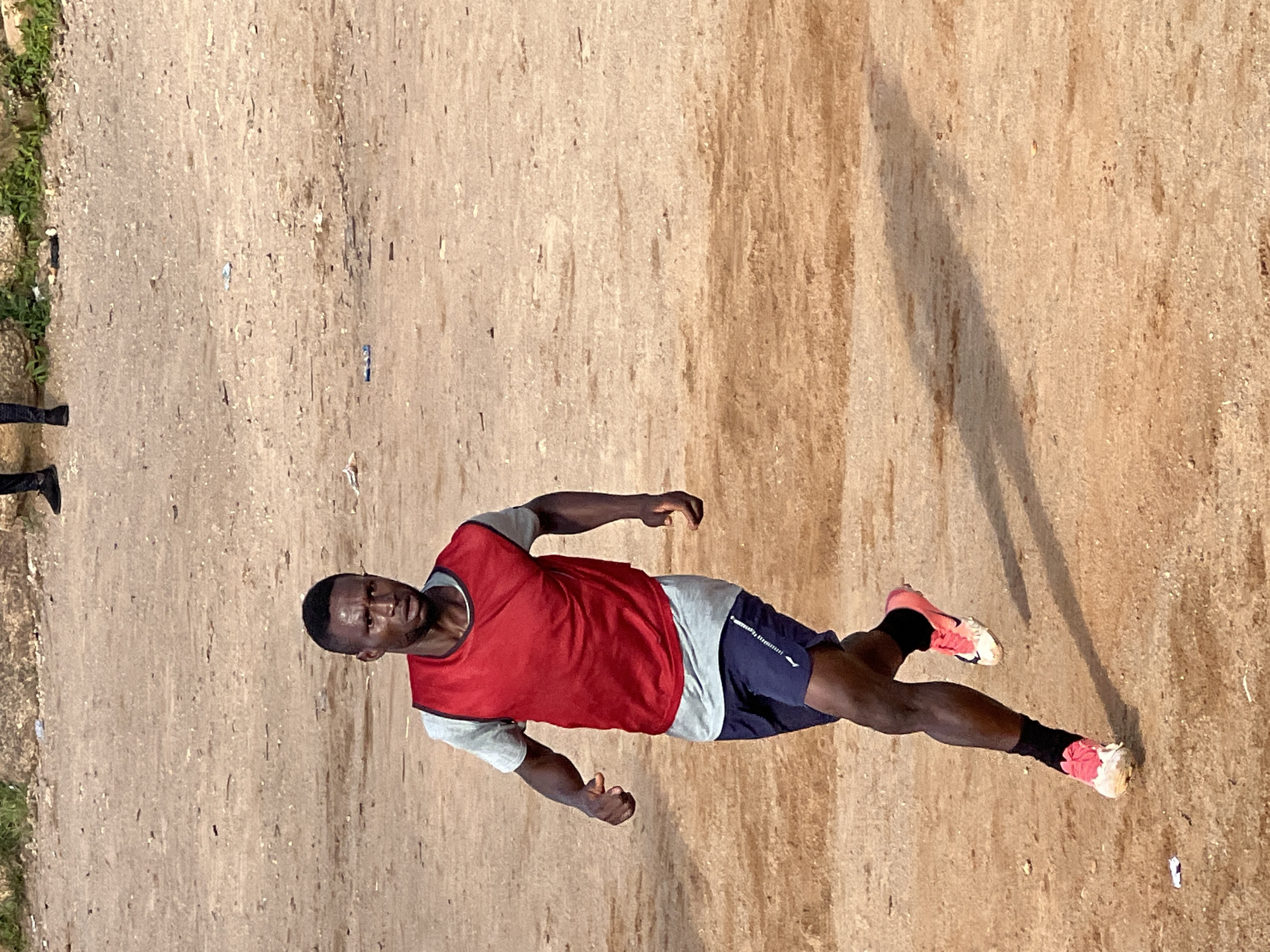 A Black male runner, wearing a grey t-shirt and a red training bib, is captured mid-stride on a dirt field. He is in the process of sprinting, with his legs in a dynamic running posture. His focus is forward, and his facial expression suggests exertion. The runner is dressed in dark blue athletic shorts and black socks with bright pink and white athletic shoes. The ground is a rough, sandy dirt surface, common in informal sports fields. The background is blurred, showing more of the same dirt terrain and a hint of vegetation at the top. There are small pieces of debris scattered on the ground. The overall impression is of an athletic training session taking place in an outdoor, unpaved area. The image is likely from Jos, Nigeria, where such informal sports grounds are prevalent.