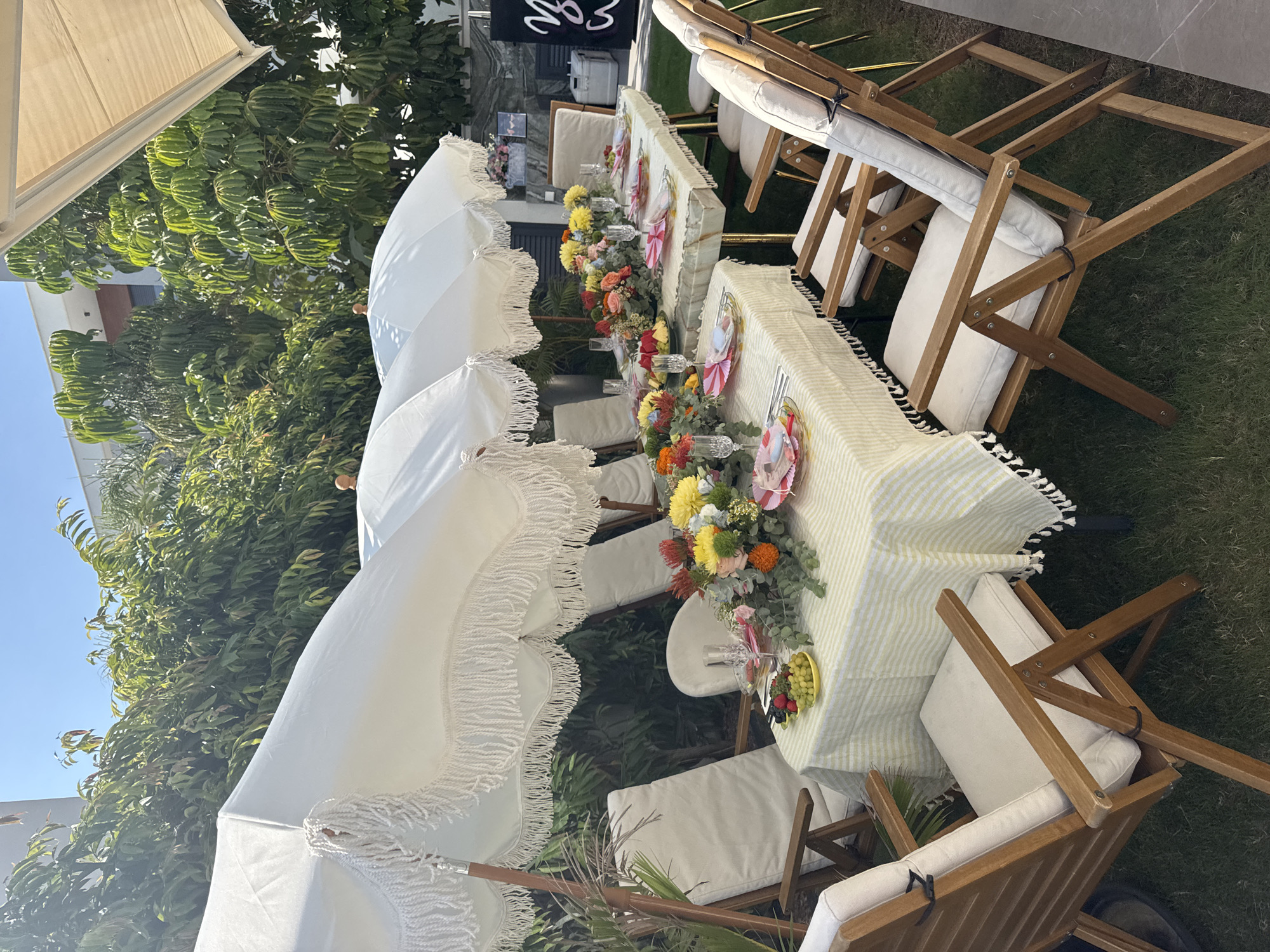 This image depicts an outdoor dining setup in Agios Tychona Community, Cyprus. Two long tables are arranged side-by-side, covered with a light yellow and white striped tablecloth that features fringed edges. Each table is meticulously set for a meal, with plates, silverware, glassware, and decorative floral arrangements. The floral centerpieces are vibrant, featuring a mix of yellow, orange, red, and purple flowers with green foliage. Small dishes of fruit, including grapes and berries, are also placed on the tables.
Surrounding the tables are several wooden chairs with white cushions. Overhead, three large white umbrellas with fringed edges provide shade. The setting appears to be in a garden or outdoor space, with lush green trees and foliage visible in the background. The lighting suggests a sunny day. To the right, a white cooler and a black sign with white lettering are partially visible. The scene suggests a prepared event, such as a party or a gathering, taking place outdoors.