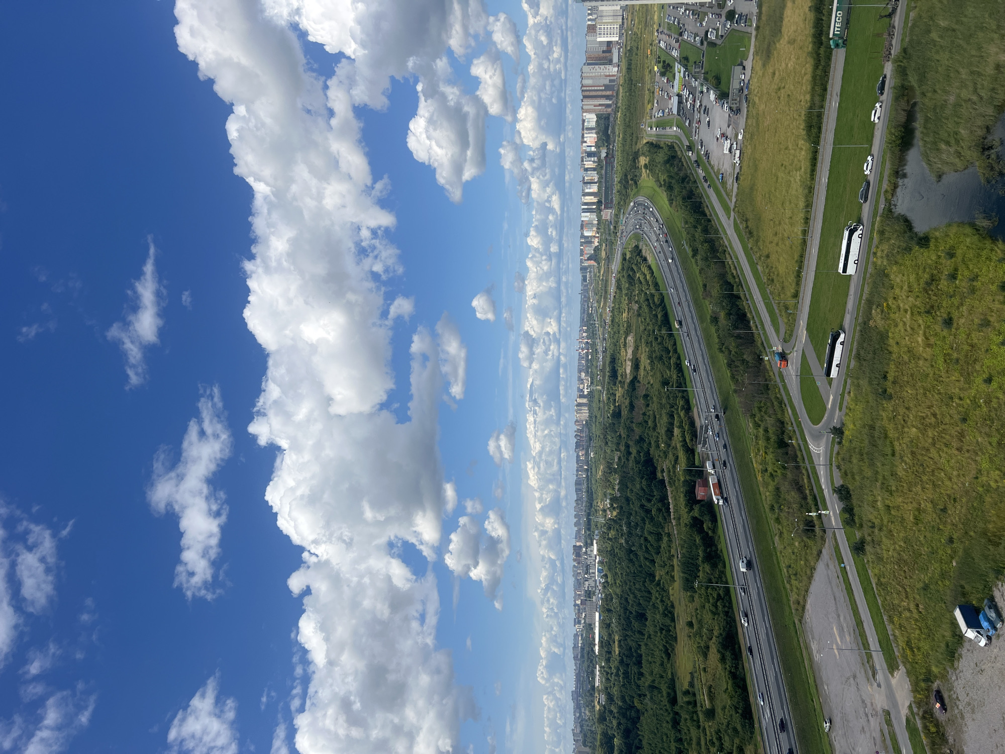An aerial view captures a vast expanse of urban landscape under a bright blue sky dotted with voluminous white clouds. A multi-lane highway curves through the scene, carrying a steady flow of traffic, including cars and trucks. Adjacent to the highway, a dense forest stretches out, giving way to a residential area composed of numerous apartment buildings. A large parking lot filled with vehicles is visible near the residential buildings. In the foreground, a body of water reflects the sky, bordered by grassy terrain. Several vehicles, including buses and cars, are present on roads in the foreground. The scene depicts a typical suburban or exurban environment with significant infrastructure and residential development.