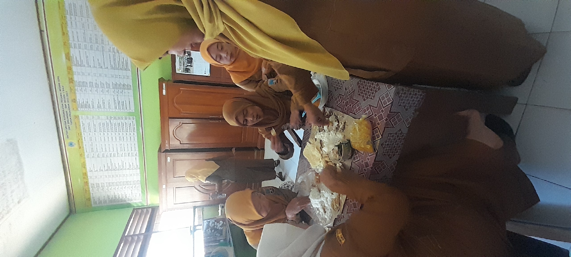 In an indoor setting, likely a classroom or meeting room, four individuals wearing brown uniforms and headscarves are gathered around a table. The table is covered with a patterned cloth and laden with food. White rice, crackers, and other food items are spread out on paper. Three of the individuals appear to be actively eating or serving themselves from the communal meal, while a fourth person in the foreground, partially visible, is also engaged with the food. In the background, there are two wooden cabinets and a green wall adorned with a large chart or poster. The lighting suggests an indoor environment, possibly during daylight hours. The scene appears to be a casual mealtime among colleagues. The presence of the chart on the wall, with Indonesian text, along with the uniforms and headscarves, suggests this location might be a school or government office in Indonesia, potentially within the Tahunan district.