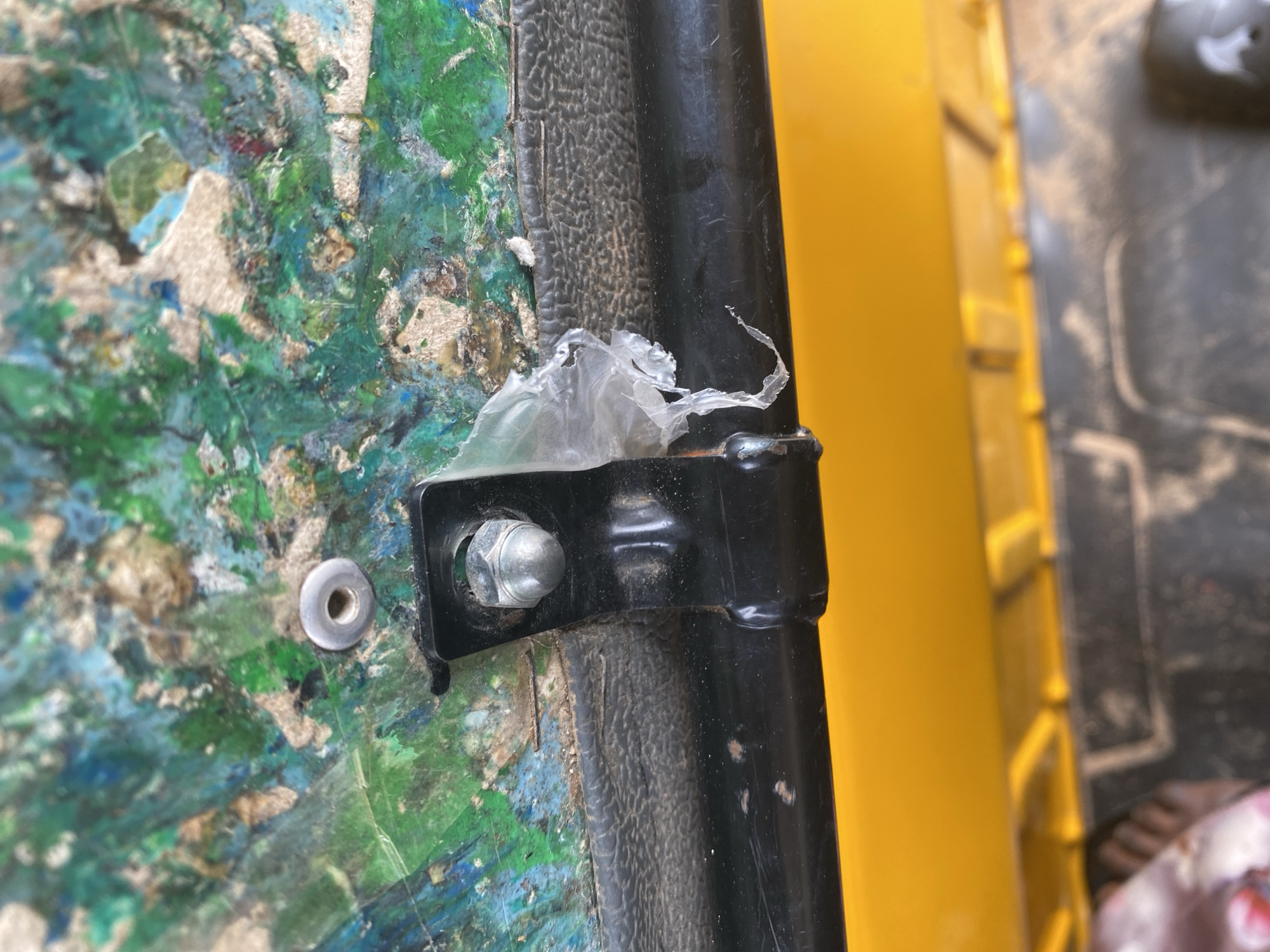 This is a close-up, eye-level shot focusing on a textured, colorful surface with a black metal bracket attached to it. The colorful surface appears to be a composite material, possibly made of layered fabrics or plastics in shades of green, blue, white, and some reddish-brown. It looks weathered and slightly damaged, with bits of material peeling or loose.
A black metal bracket is bolted onto this surface, holding a cylindrical black rod in place. The bolt itself is visible, a hexagonal nut with a visible threaded shaft. There's a piece of clear, crinkled plastic caught between the colorful surface and the black bracket, adding a layer of detail and suggesting a less-than-pristine condition. To the left of the bracket, there's a round, metallic object, likely a washer or a rivet, embedded in the colorful material.
The background is a bright, solid yellow, suggesting either a painted surface or a yellow object. To the far right, a darker, textured surface is partially visible, with some reddish and white elements that might be part of another object or debris.
The scene appears to be an outdoor setting, given the texture and possible weathering of the colorful material. No people are visible. The lighting is bright, casting distinct shadows and highlighting the textures. The overall impression is one of a functional, perhaps utilitarian, object or structure that has seen some use and is exposed to the elements. The location context provided (Jalingo, Nigeria) doesn't offer specific visual cues within the image itself, but suggests a warm, possibly sunny, environment. There is no visible text in the image.
