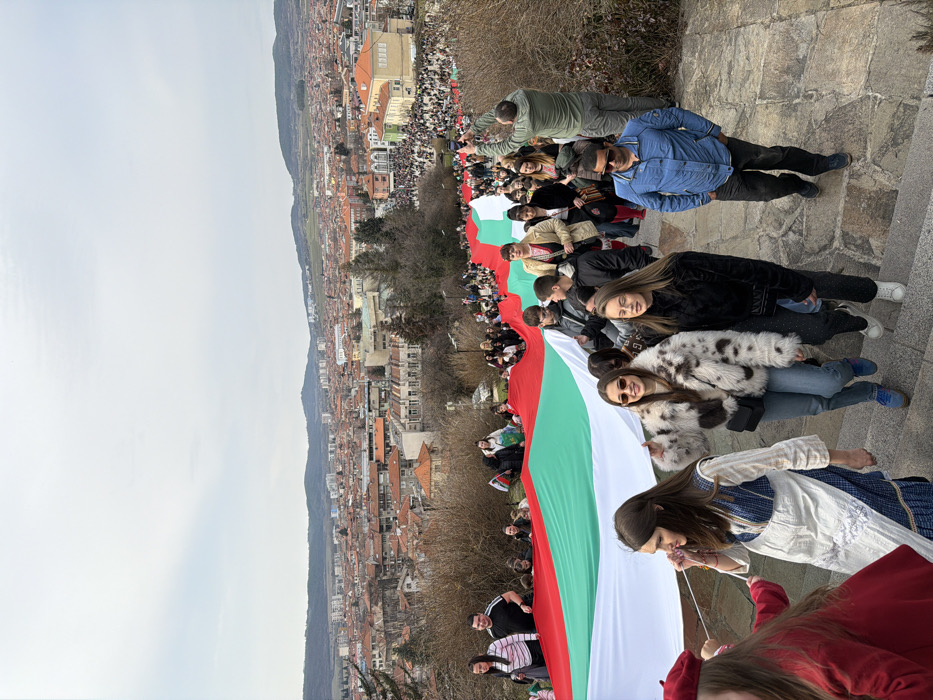 The image captures a large procession in Panagyurishte, Bulgaria, from an elevated vantage point. A very long and wide Bulgarian national flag, featuring horizontal white, green, and red stripes, is being carried by numerous individuals along an outdoor stone staircase.
In the immediate foreground, a young woman in a white and blue traditional-style dress is visible on the left. Adjacent to her, a young woman wearing a fur-patterned jacket, jeans, and sunglasses smiles towards the camera, accompanied by another young woman in a dark top and trousers. To their right, a man in a blue jacket and sunglasses walks on the stairs. Further back in the procession, a man in a green jacket is seen holding a smartphone aloft, appearing to record the event. The crowd extends into the distance, actively holding the expansive flag as it undulates down the staircase.
The city of Panagyurishte forms the background, characterized by a dense arrangement of buildings, primarily residential structures with red-tiled roofs, and some taller, more modern constructions on the outskirts. Rolling, treed hills define the distant horizon under an overcast sky. The stone steps are flanked by bare deciduous trees and shrubs, suggesting a cooler season. A large congregation of people is visible in a public square or park area within the city, further down the procession path.