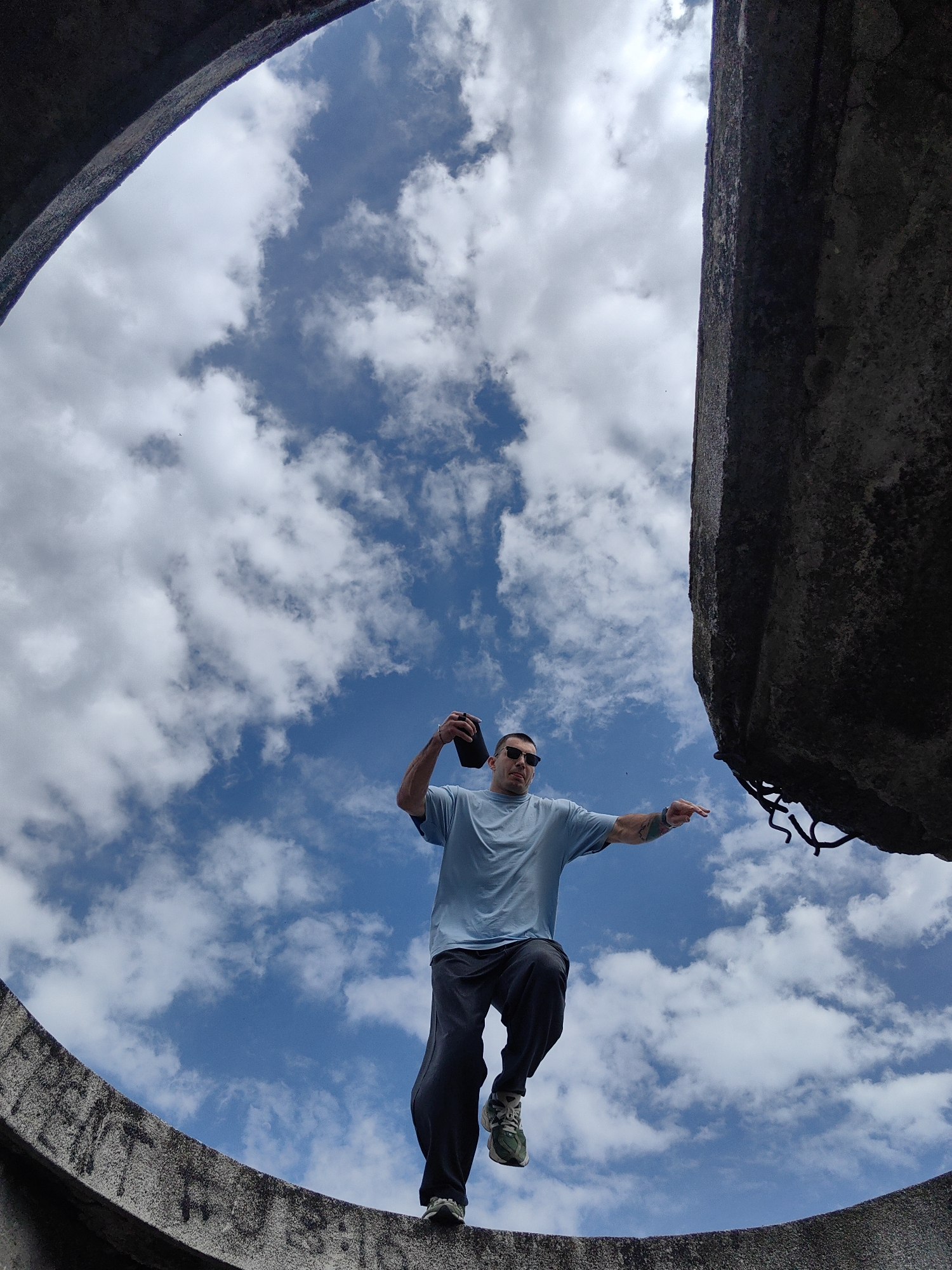 A man is standing atop a concrete structure against a bright, cloudy sky, seemingly within a circular opening. He is holding a dark object in one hand and pointing with the other, while wearing sunglasses. The location is Varna, Bulgaria, inside the ruins of the unfinished monument. The monument is on a hilltop overlooking the city. The man is positioned so that parts of the concrete structure frame the sky, suggesting he is inside the monument.