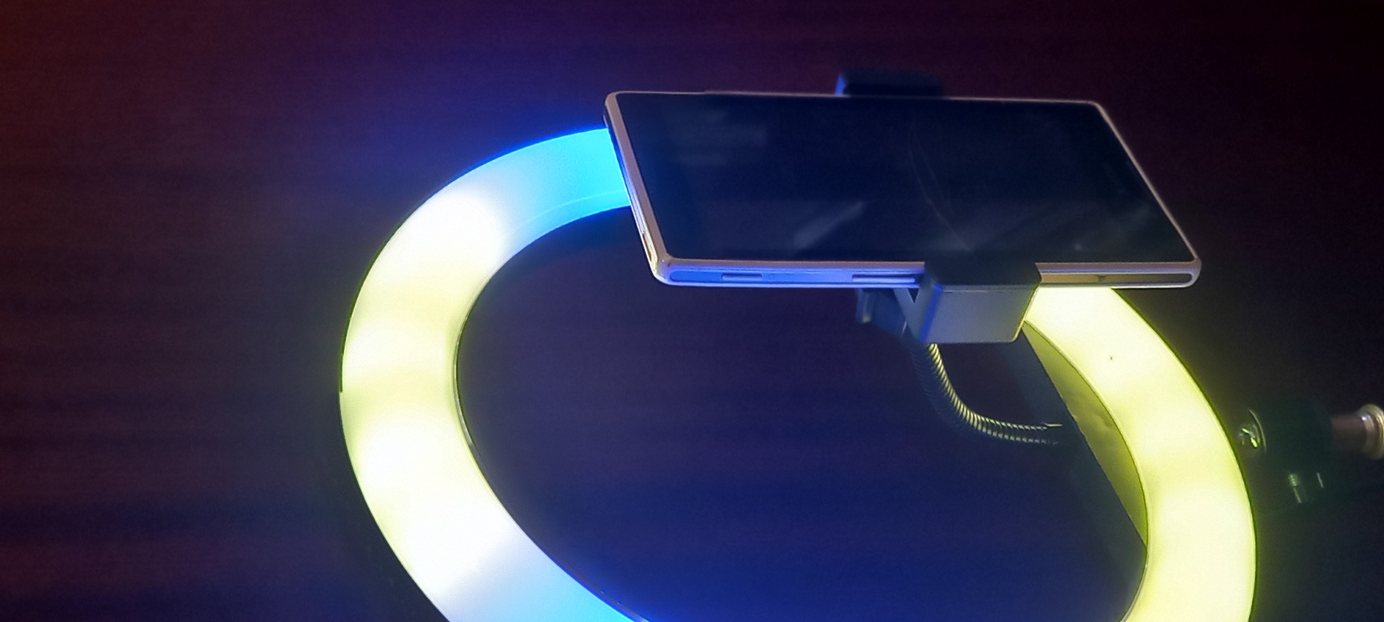 The image displays a close-up, angled view of a smartphone mounted within a circular ring light against a dark, indistinct background. The scene suggests an indoor setting, likely in Kryvyi Rih, Ukraine, with low ambient light, possibly during nighttime.
The primary subjects are a modern smartphone, held horizontally by a dark grey or black plastic mount, and the illuminated ring light. The phone's screen is dark, showing faint reflections or minor smudges, with control buttons visible on its side. The ring light emits a vivid color gradient, transitioning from a cool, bright light blue on the left side to a warm, soft yellow or off-white on the right. This gradient provides the main source of illumination, casting a glow that highlights the phone and subtly illuminates the immediate surroundings.
A flexible, dark, ribbed gooseneck arm is partially visible, connecting the phone holder to the ring light assembly. This setup is typical for activities requiring controlled front-facing lighting, such as video calls, live streaming, vlogging, or taking selfies and portraits. The dark background, rendered in deep blues and purples, further emphasizes the luminous quality of the ring light. There is no visible text in the image, nor are there any people or specific activities depicted beyond the arrangement of the technological equipment.