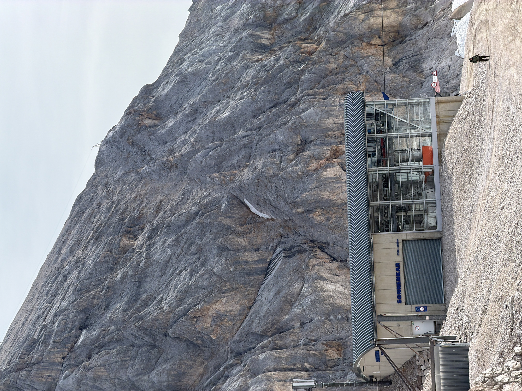 The image shows the "Sonnenkar" cable car station at the Zugspitze mountain near Garmisch-Partenkirchen, Germany, at an altitude of 2300-2800 meters. A large, modern building with a corrugated metal roof and glass facade houses the cable car machinery. The station is situated on a steep, rocky mountainside, with sparse vegetation and patches of snow visible. The ground in front of the station is covered in gravel and dirt. A single person, dressed in dark clothing, is walking away from the station on a dirt path. The sky is clear and bright.