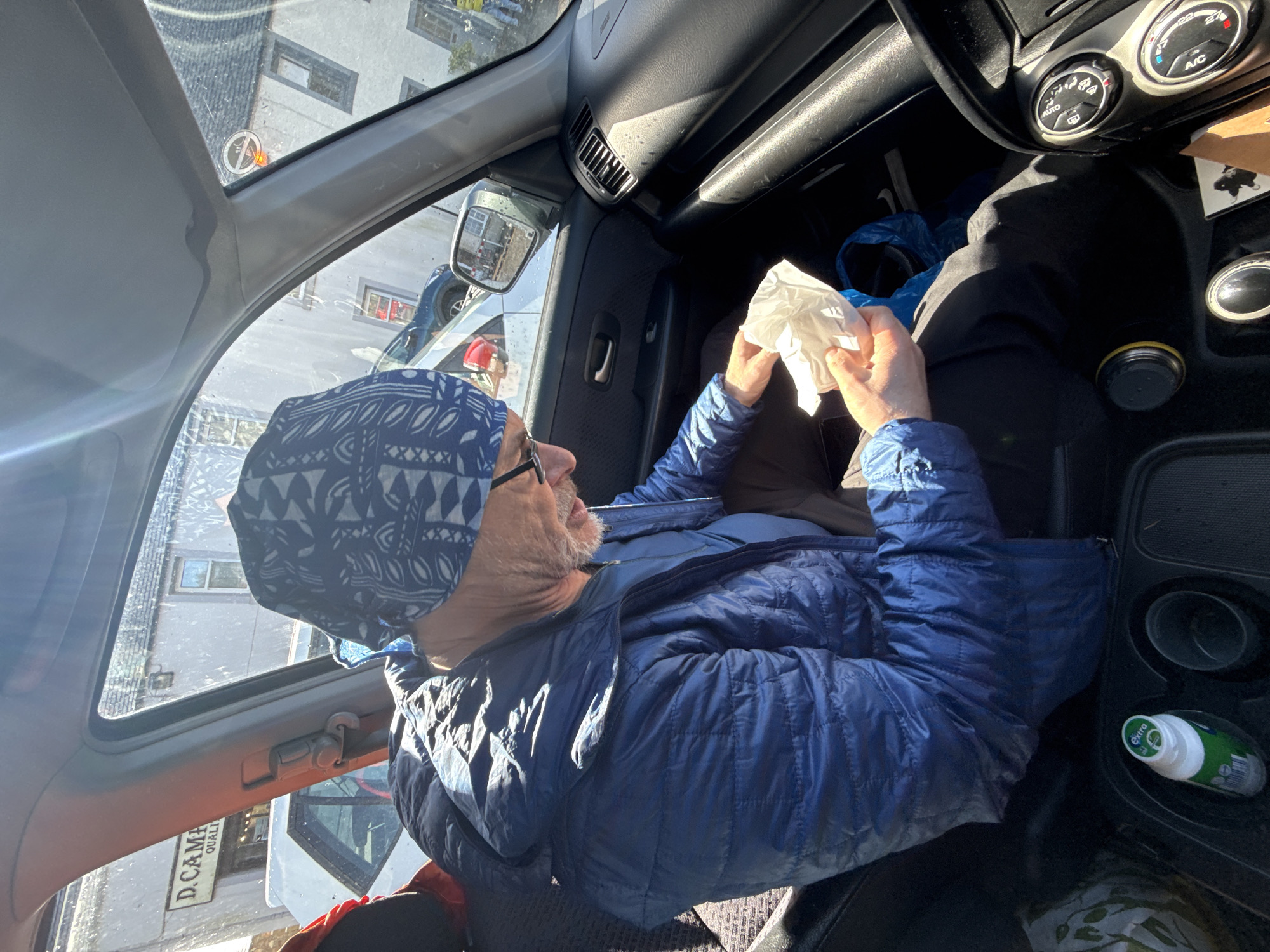 An adult male is seated in the front passenger seat of a vehicle, partially illuminated by bright sunlight. He wears a blue quilted jacket, a patterned blue and white head covering, and eyeglasses. He has a grey beard and is engaged with a crumpled white wrapper or tissue held in both hands. The vehicle's interior features a dark dashboard, a partially visible center console with a green "Extra" brand bottle in a cup holder, and climate control dials. Outside the passenger window, an urban street scene is visible. A white car is parked or driving in front of a building with a prominent sign reading "D.CAMPBELL QUALITY." Other light-colored, multi-story buildings with grey pitched roofs line the street, indicating an established town or city environment. A "no smoking" decal is affixed to the upper right portion of the interior windshield. The scene occurs in the city of Unknown, Unknown.