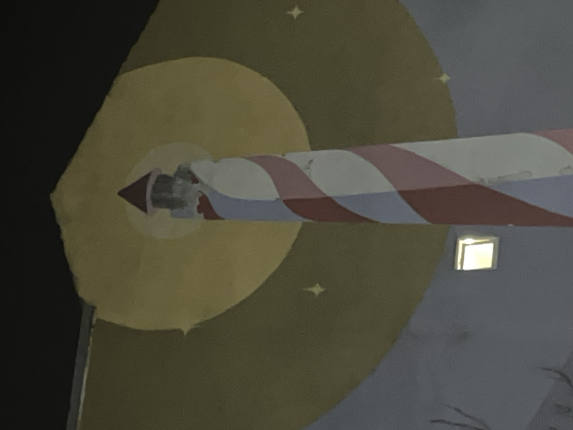 A cropped indoor shot of a wall mural depicts a stylized lighthouse with a red and white striped cylindrical body. The lighthouse emerges from a yellow halo that appears to represent the moon or a light source. The background features a dark, star-speckled sky. A window with a lit interior is also visible. There is no verifiable evidence that this mural is in Budva, Montenegro.