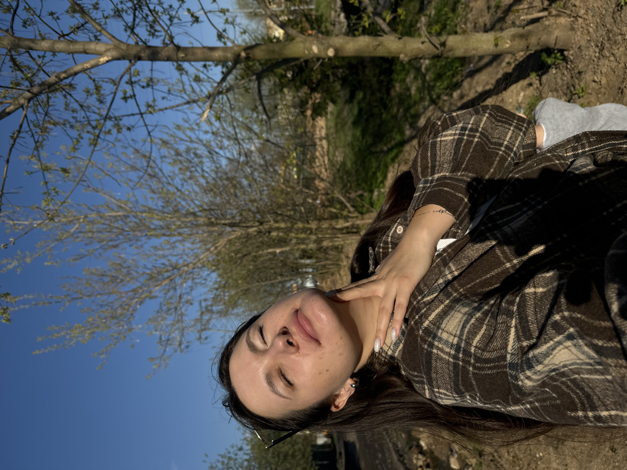 A woman with dark hair is lying on the ground outdoors, possibly in Akulovo, Russia, given the broader location of Gp Kubinka. She is wearing a brown and tan plaid shirt and grey socks, resting her hand on her neck. The background consists of a blue sky and trees with budding leaves. The ground appears to be soil or dirt.