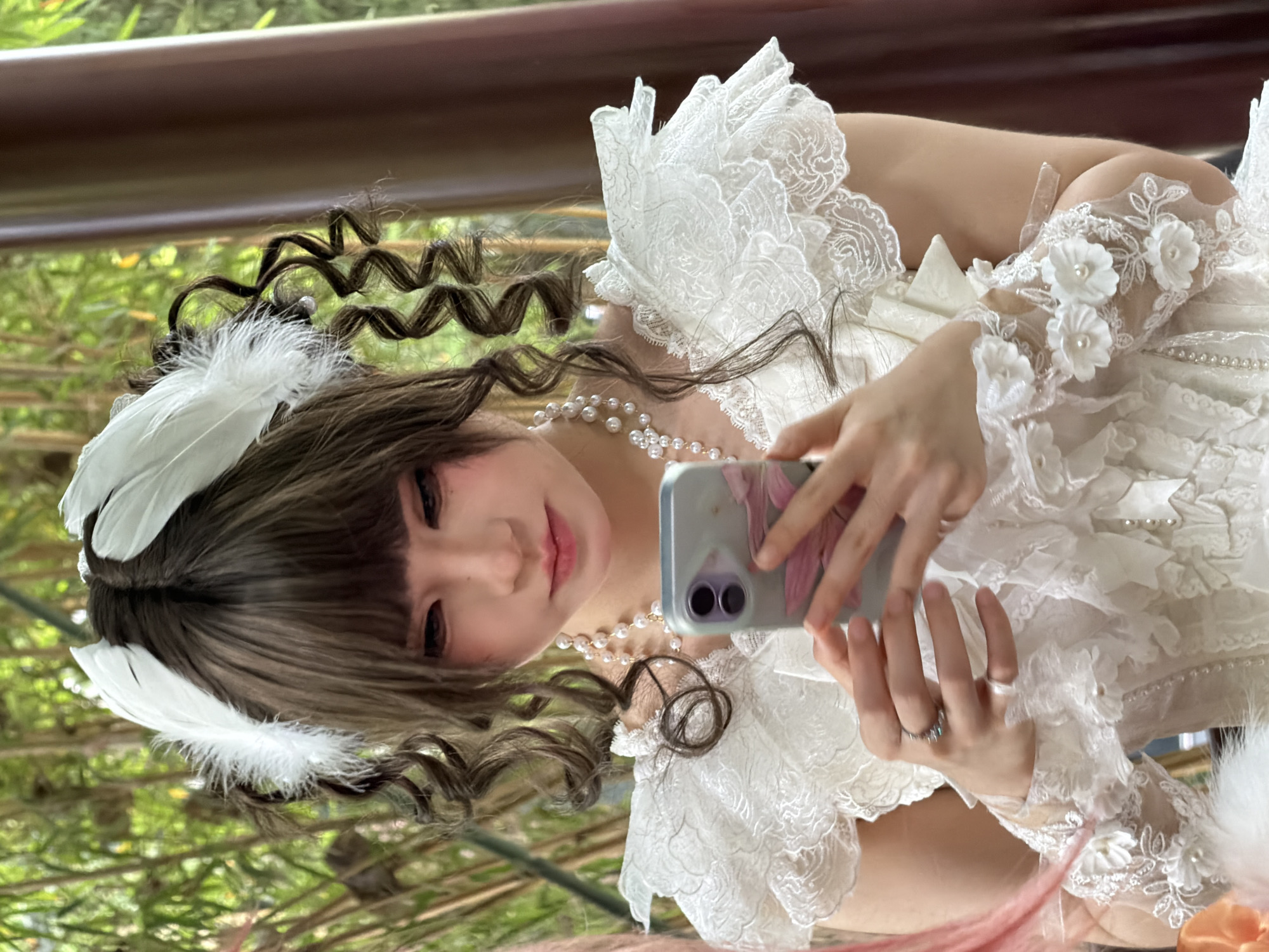 A young Asian woman with long, dark, curly hair, adorned with white feathers, is holding a smartphone in her right hand and looking at it. She is wearing a white, ruffled dress with lace and pearl embellishments. A pearl necklace is visible around her neck. The background appears to be outdoors with green foliage and a bamboo-like structure. The setting is likely a garden or a park within Yuzhong District, China.