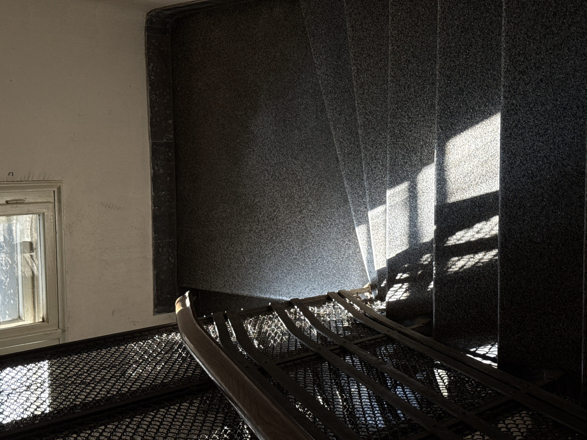 The image shows a stairwell in an old building. The walls are finished in a dark granite or stone. A window is visible on the left. The stairs have a metal mesh surface. The staircase is lit by sunlight casting shadows across the steps and the adjacent wall. The location is Belgrade, Serbia.
