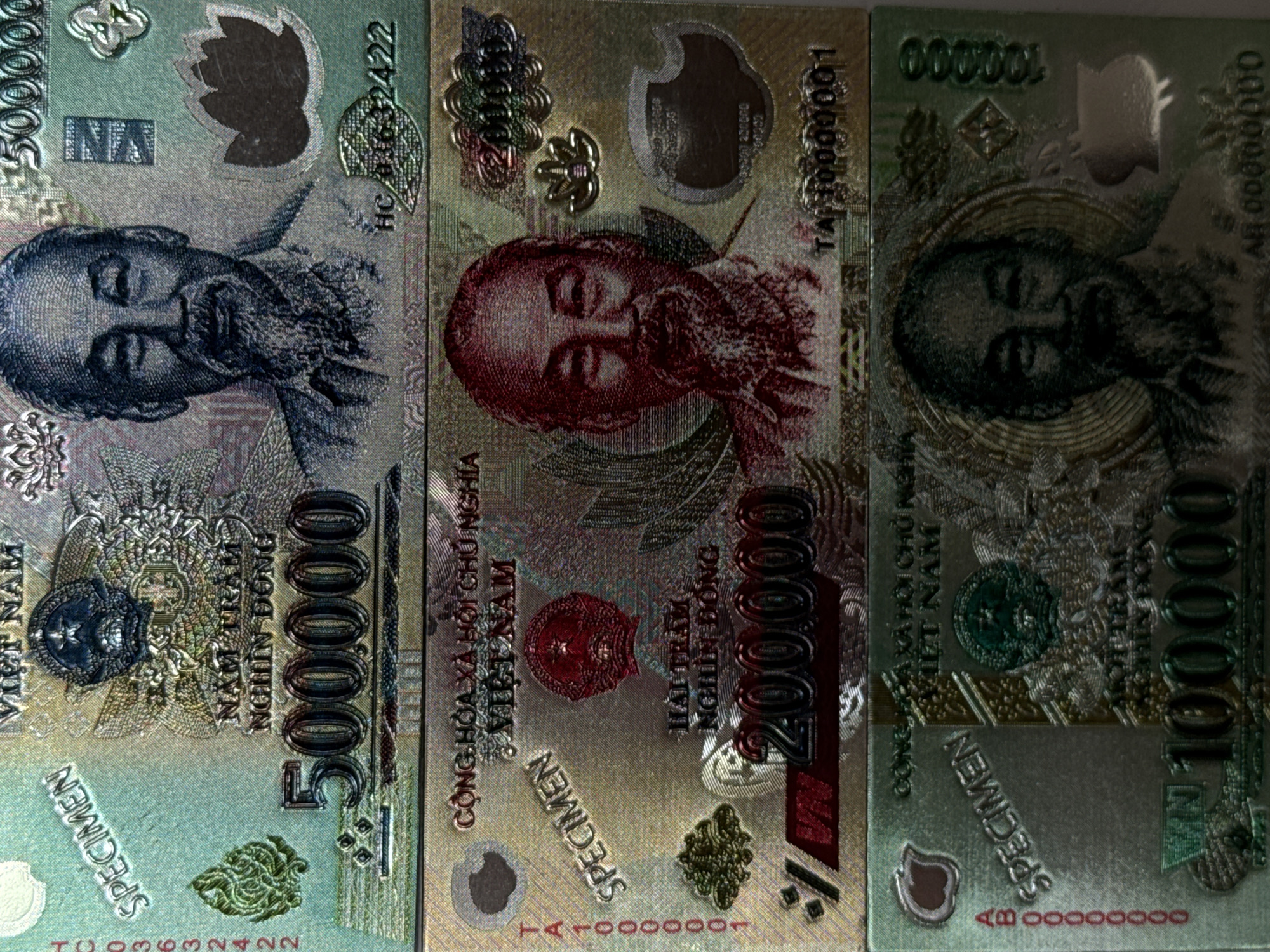 The image displays three Vietnamese Dong banknotes. The banknotes are laid out side-by-side. The denominations visible are 500,000, 200,000, and 100,000 Dong. Each features a portrait of Ho Chi Minh. The banknotes are labeled "SPECIMEN." Serial numbers are visible on each. The backdrop appears to be a flat surface, possibly a table. There is no indication of Izhevsk, Russia in this image.