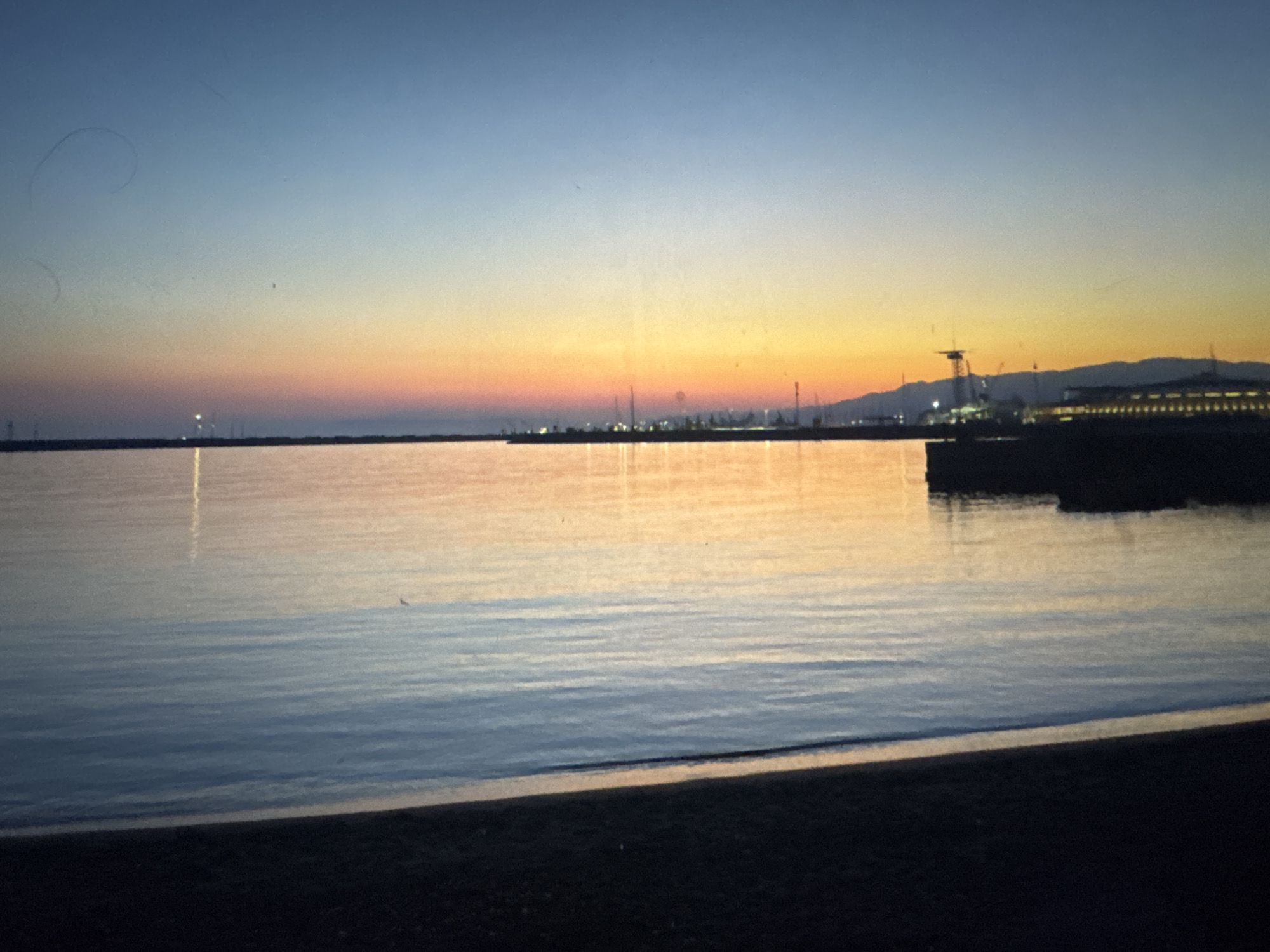The image shows a serene coastal scene at dusk. The sky transitions from a deep blue at the top to hues of orange and pink near the horizon, indicating sunset or sunrise. The calm water reflects the colors of the sky and the distant lights of a city or harbor. Silhouettes of structures, possibly cranes and buildings, are visible along the coastline. A narrow strip of dark sand or beach occupies the foreground. The overall atmosphere is tranquil and peaceful. The image does not contain any elements that would suggest it is located in Porrentruy, Switzerland, as Porrentruy is an inland town.