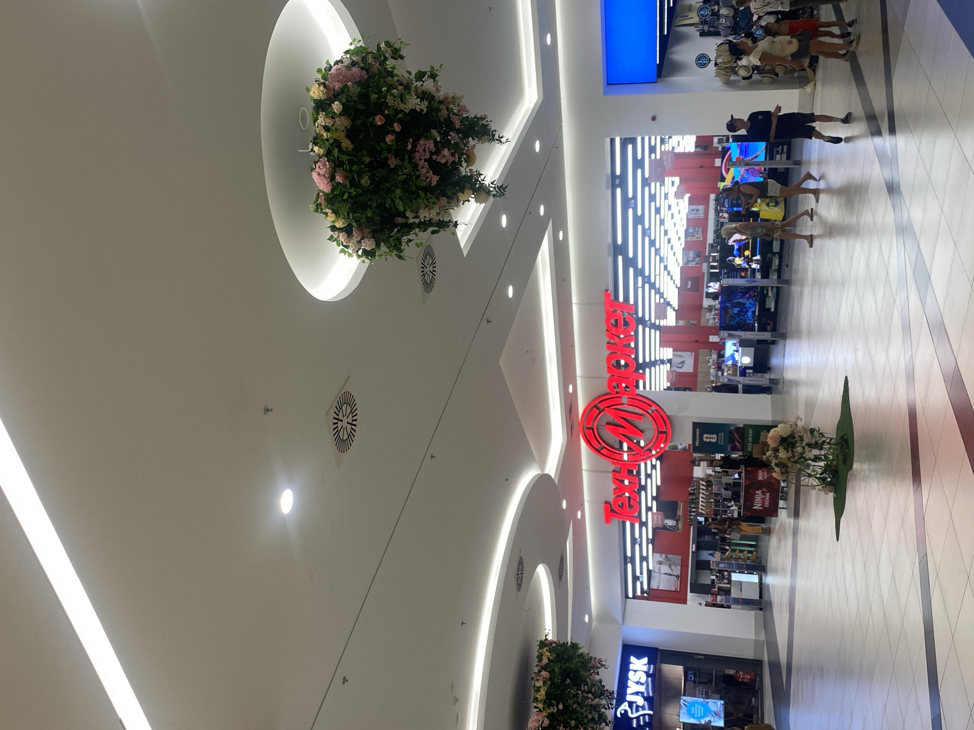 This is a medium-angle, full shot taken inside a brightly lit shopping mall, likely in Burgas, Bulgaria. The ceiling is white and features a series of modern, curved light fixtures and circular air vents. Hanging from the ceiling are two large, spherical floral arrangements, one pink and white with greenery, and the other predominantly green with hints of pink and yellow.
In the center of the image, the prominent red neon sign for "Техномаркет" (Technomarket) is visible, illuminated against a backdrop of white. In front of Technomarket, there are display stands with televisions and other electronic equipment. To the left of Technomarket is a store with a blue and white sign reading "JYSK".
The mall floor is tiled with a pattern of light grey and white squares. Several people are walking through the mall. On the left, a person is entering the JYSK store. In the mid-ground, a group of people, including two women walking together and a person in a dark t-shirt and shorts walking away from the camera, are moving through the mall. Further to the right, a family or group of people, including adults and children, are browsing merchandise.
In the foreground, a small floral arrangement with pink and white flowers is placed on a patch of artificial grass. The overall impression is a modern, clean, and well-maintained shopping environment during the daytime.