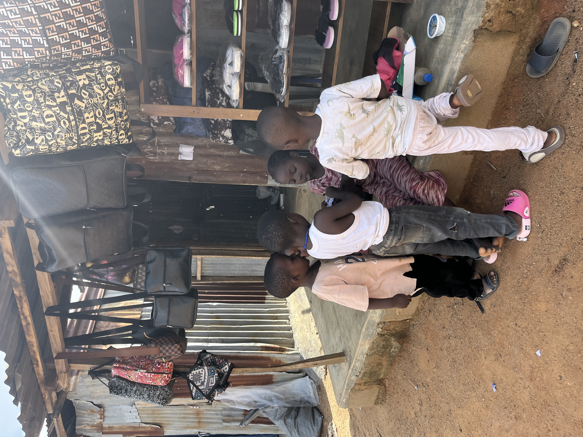 The image shows a group of young children interacting in an outdoor market setting. In the foreground, four children are gathered. One child, wearing a white tank top and jeans, leans in towards another child. The second child, in a light pink t-shirt, is partially obscured. A third child, dressed in a patterned long-sleeved shirt and pink pants, is lying down. A fourth child, also in a white long-sleeved shirt with a pattern of green reptiles and light pink pants, is lying next to the third child. The children appear to be engaged in a close interaction, possibly playing or talking.
In the background, a stall displays various bags and merchandise on shelves. There are several duffel bags with prominent logos, including one with a repeating "F" pattern and another with letters and numbers. Other bags and items are also visible on the shelves. The stall appears to be made of wood and corrugated metal, suggesting a rustic or informal marketplace.
To the right of the children and the stall, there are some shoes and a bottle of water on the ground. A blue slipper is visible on the ground.
The lighting suggests it is daytime, possibly late morning or early afternoon, with bright sunlight casting shadows. The ground is covered with dirt and gravel, and the overall scene depicts a typical day in a marketplace in Jos, Nigeria. The children's expressions are not clearly visible, but their close proximity suggests familiarity and comfort with each other. There is no visible text in the image, other than patterns on the bags which are not clearly legible.