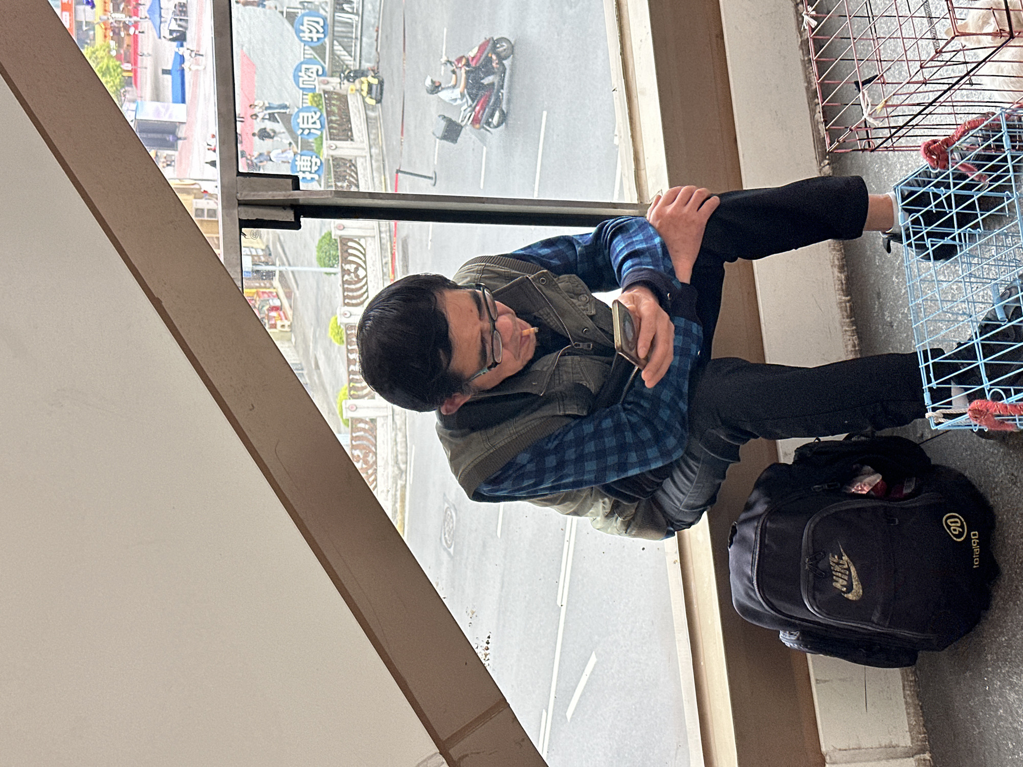 The image depicts a man sitting indoors next to a window overlooking an urban scene. The man, wearing glasses, a blue plaid shirt, and a vest, is holding a cell phone and has what appears to be a cigarette in his mouth. He is sitting with his legs crossed and close to a black Nike backpack and a blue wire cage. Outside the window, there is a street with moving vehicles and pedestrians. In the background, the image shows signs written in Chinese, suggesting the location is in Jiulongpo District, China.