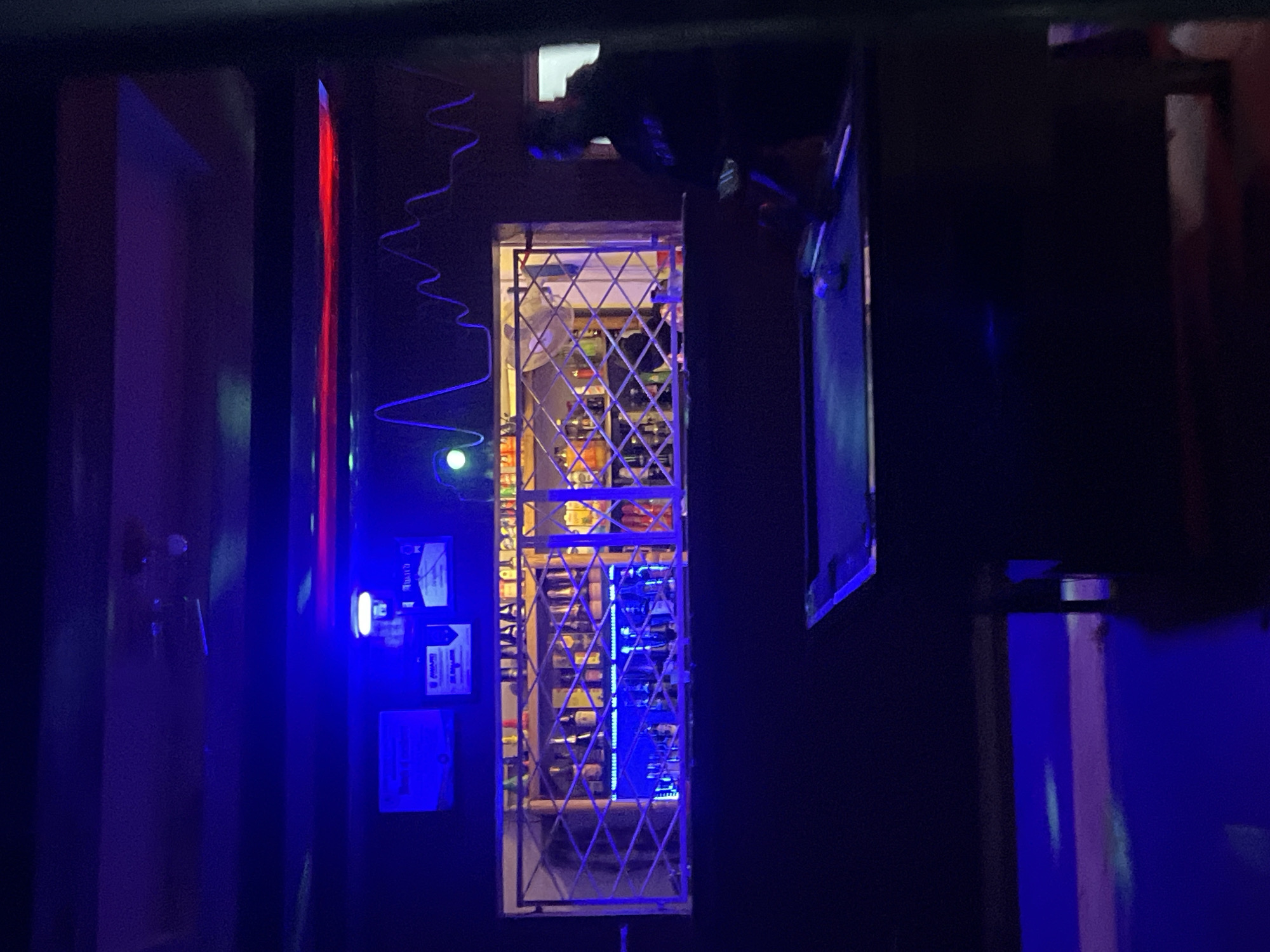 The image depicts the interior of a dimly lit bar, likely in Durumi, Nigeria, illuminated by vibrant blue and red neon lights. A bar counter dominates the foreground, with a pool table partially visible in the lower right corner. Behind the counter, shelves stocked with numerous bottles are visible through a metal grate, suggesting a well-supplied establishment. Several framed awards or certificates hang on the wall above the bar. The overall ambiance is that of a lively nightlife venue.