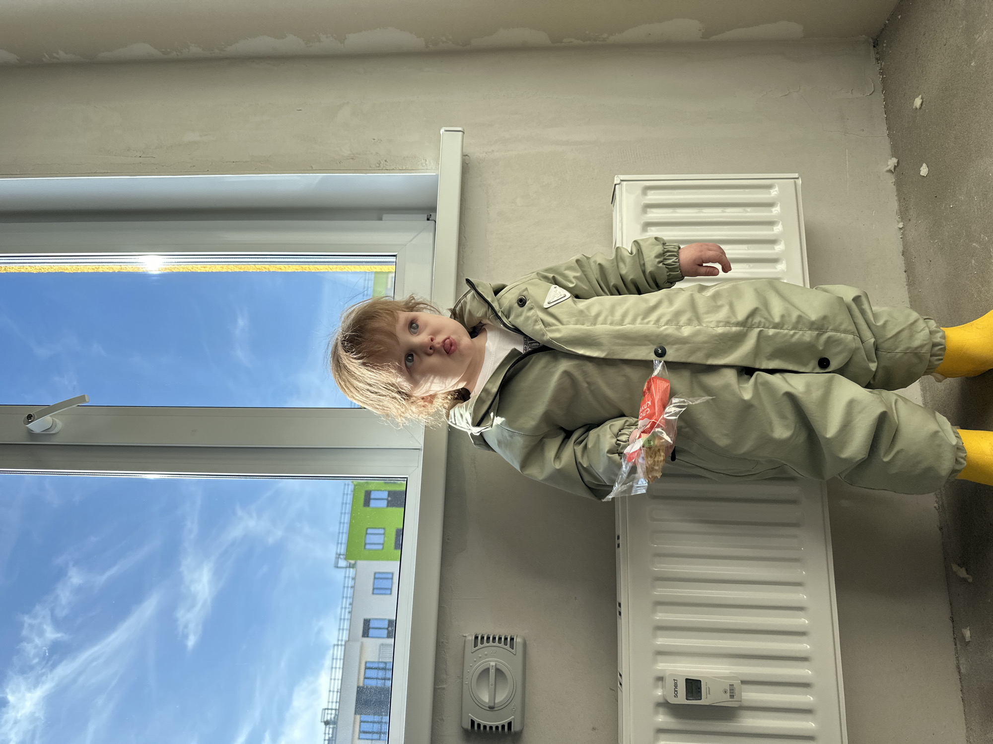 A young child stands in an unfurnished room within a building under construction or renovation in Pervomaysky, Russia. The child, with light hair, wears a light olive-green full-body jumpsuit and bright yellow boots. They hold a transparent plastic bag containing crackers, with red text, in their right hand, and have their tongue slightly extended. Behind the child, a white-framed window provides a view of a clear blue sky with wispy clouds and distant multi-story apartment buildings, one featuring a prominent bright green facade. Below the window, a white panel radiator is mounted, equipped with a "Sanext" brand heat meter. To the left of the radiator, a grey wall-mounted ventilation unit with a circular control dial is present. The interior walls are unfinished, displaying patchy, unpainted surfaces, and the floor is a bare concrete slab.