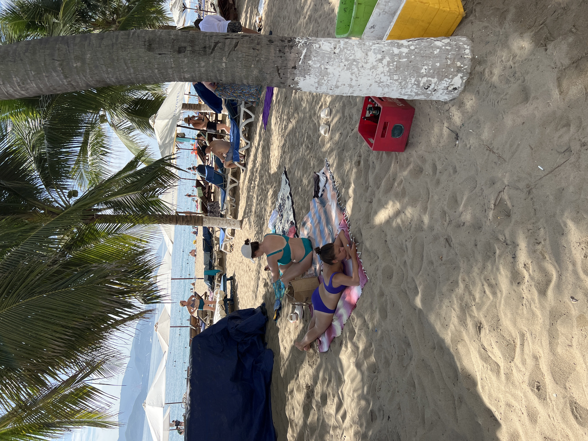 The image depicts a beach scene in Nha Trang, Vietnam, on a sunny day. The foreground is dominated by sandy beach with footprints and shadows cast by palm trees. Two women are lying on towels. One, in a purple bikini, is prone on her stomach, looking at her phone. Another, in a teal bikini, sits up on a towel with her back to the viewer, appearing to be packing a bag. In the background, a row of white beach umbrellas covers lounge chairs. The ocean is visible beyond the chairs, with a few people swimming and sunbathers. Several palm trees line the beach, with one prominently featured in the right foreground, its trunk painted white at the base. A red crate filled with bottles and two plastic bins (one green, one yellow) are on the sand near the palm tree. The overall atmosphere is relaxed and typical of a tropical beach resort.