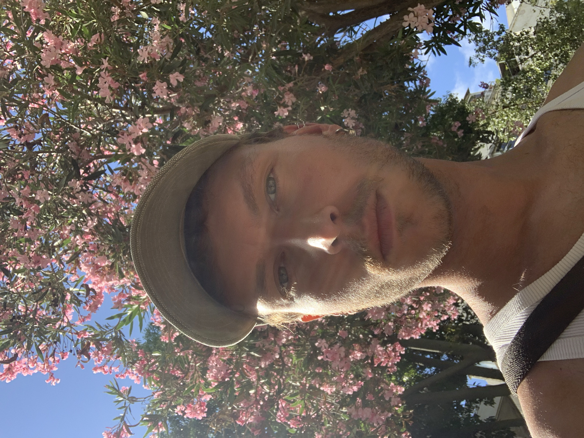 A selfie of a fair-skinned man with blue eyes and short brown hair wearing a hat and white tank top with a dark shoulder strap. He has a light beard. Behind him is a flowering tree with pink blossoms and green leaves. The sky is blue and partially visible through the branches. The location is possibly Tel-Aviv, Israel.
