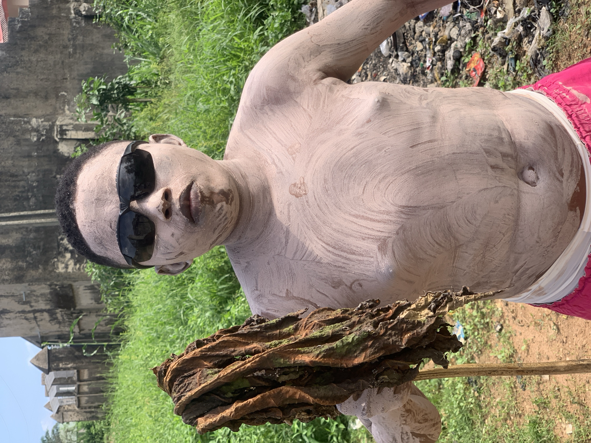 A person covered in light brown mud stands in an outdoor environment. The person is wearing dark sunglasses and a white band around their waist, with pink fabric below it. They are holding a large, dry, brown leaf on a wooden stick. In the background, there is lush green vegetation, several old, weathered buildings with pointed roofs, and a pile of debris. The ground is a mixture of dirt and scattered refuse. The sky is clear and blue. The location appears to be within a built-up area, likely in the city of Ikorodu, Nigeria, given the distinctive architecture of the buildings.