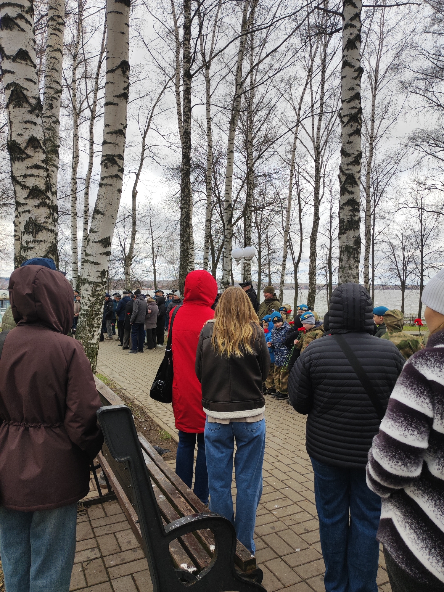 A group of people stands in a line on a paved walkway next to a park in Votkinsk, Russia. The line extends into the distance, comprised of both adults and children. Some individuals carry red flowers. Several trees line the walkway on either side, mostly birch trees with distinctive black and white bark. Beyond the park, a body of water is visible under an overcast sky. In the foreground, a brown hooded jacket and a red hooded jacket are seen. A park bench is to the left.