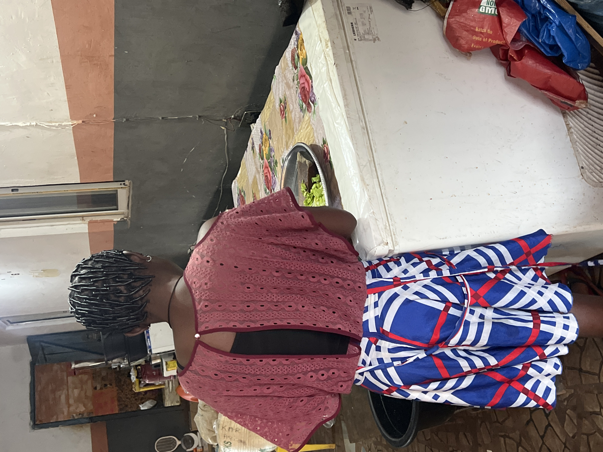 A medium shot captures a Black woman from the rear, standing in what appears to be a kitchen or food preparation area in Nkpor, Nigeria. She has her hair in braided cornrows and wears a dark-colored tank top beneath a loose-fitting, short-sleeved, open-back pink blouse with a circular cut-out at the back, embellished with eyelet patterns. Her skirt is a vibrant A-line design featuring a geometric pattern of blue, white, and red stripes and squares.
The woman is positioned in front of a white chest freezer, which is covered with a cloth featuring a floral print of roses and other flowers. On the freezer, a metal bowl containing chopped green vegetables, possibly lettuce, rests on a wooden board. To the left of the woman, a large black plastic basin is visible. The background reveals a utilitarian space with cinder block walls painted in sections of dark gray, white, and a rusty orange. A doorway with a dark door stands open in the left background, showing stacked items and a gas cylinder. To the right of the freezer, several plastic bags, predominantly red and blue, are piled. The floor is tiled with a mosaic pattern. The overall scene suggests a domestic or small-scale commercial food operation.