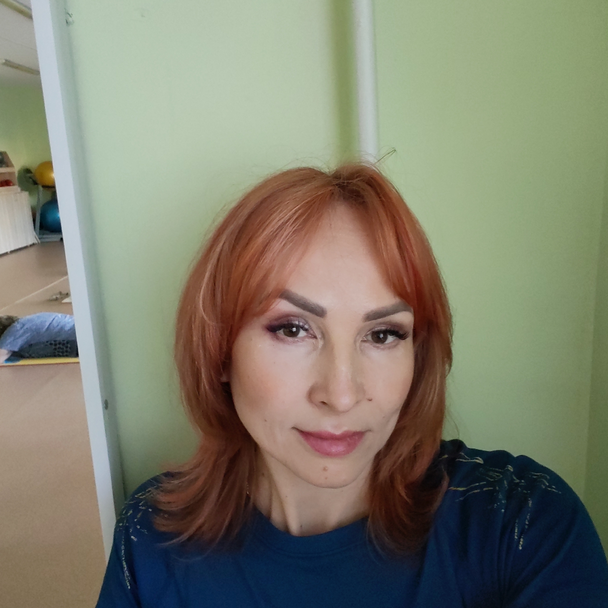 A woman with reddish-orange hair poses for a selfie. She is wearing a dark blue shirt with a subtle gold pattern on the shoulder. She has fair skin, brown eyes, and wears makeup, including eye shadow and lipstick. She appears to be indoors, standing in front of a light green wall. A white door frame is visible to the left, revealing a room with a beige floor, white furniture, and exercise equipment such as an exercise ball. The location is Chaikovsky, Russia. A person is lying on the floor in the background.