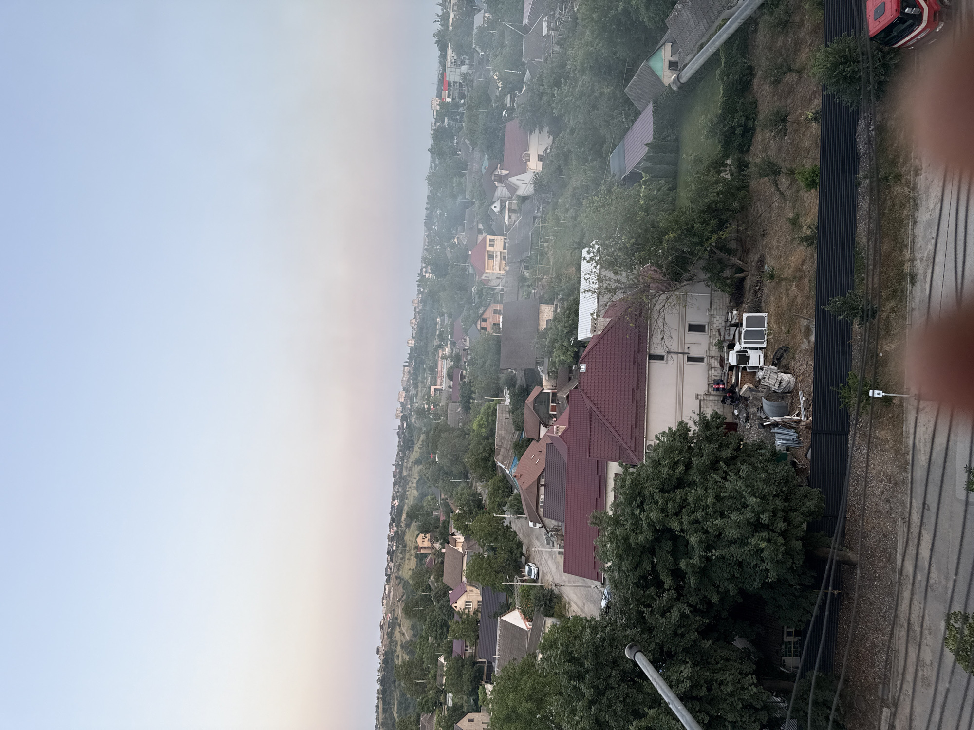 This is a vertical, aerial photograph of a residential neighborhood in Cahul, Moldova. The scene is dominated by numerous houses with red and brown tile roofs, interspersed with lush green trees. A road winds through the neighborhood, with a few cars visible. In the foreground, a pile of scrap metal and debris is visible next to a large tree and a fence. Further down the road, a red truck is parked. The sky is overcast, with a gradient of light blue to pale yellow, suggesting it may be early morning or late afternoon. The overall impression is one of a densely populated, somewhat cluttered, semi-rural or suburban environment. There is no visible text in the image.