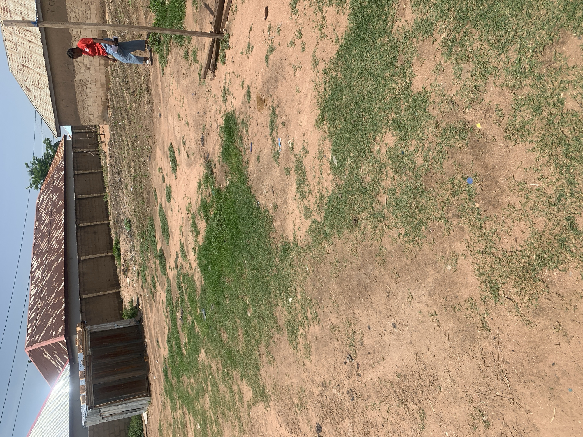 The image shows an outdoor scene with a person standing near a building. The person, a young Black individual wearing a red t-shirt and blue jeans, is looking down at a tablet or book. They are positioned on a dirt and grass-covered ground, with a weathered building to their left. The building has a corrugated metal roof that appears aged and discolored.
To the right of the person, the ground is a mix of dry soil and patches of green grass. Scattered across the ground are small pieces of colored paper, including yellow and blue. In the background, part of another building with a similar corrugated roof is visible, as well as a tall, thin wooden pole. The overall scene suggests a somewhat unkempt or undeveloped area.
The lighting indicates it is daytime, likely with bright sunlight casting shadows, although the exact time of day is difficult to determine. The weather appears to be clear and possibly warm. The location is identified as Jauro Yinu, Nigeria, which provides context for the architectural style and the arid/semi-arid environment suggested by the ground cover. There is no discernible text in the image. The activity appears to be a moment of quiet observation or study by the individual.