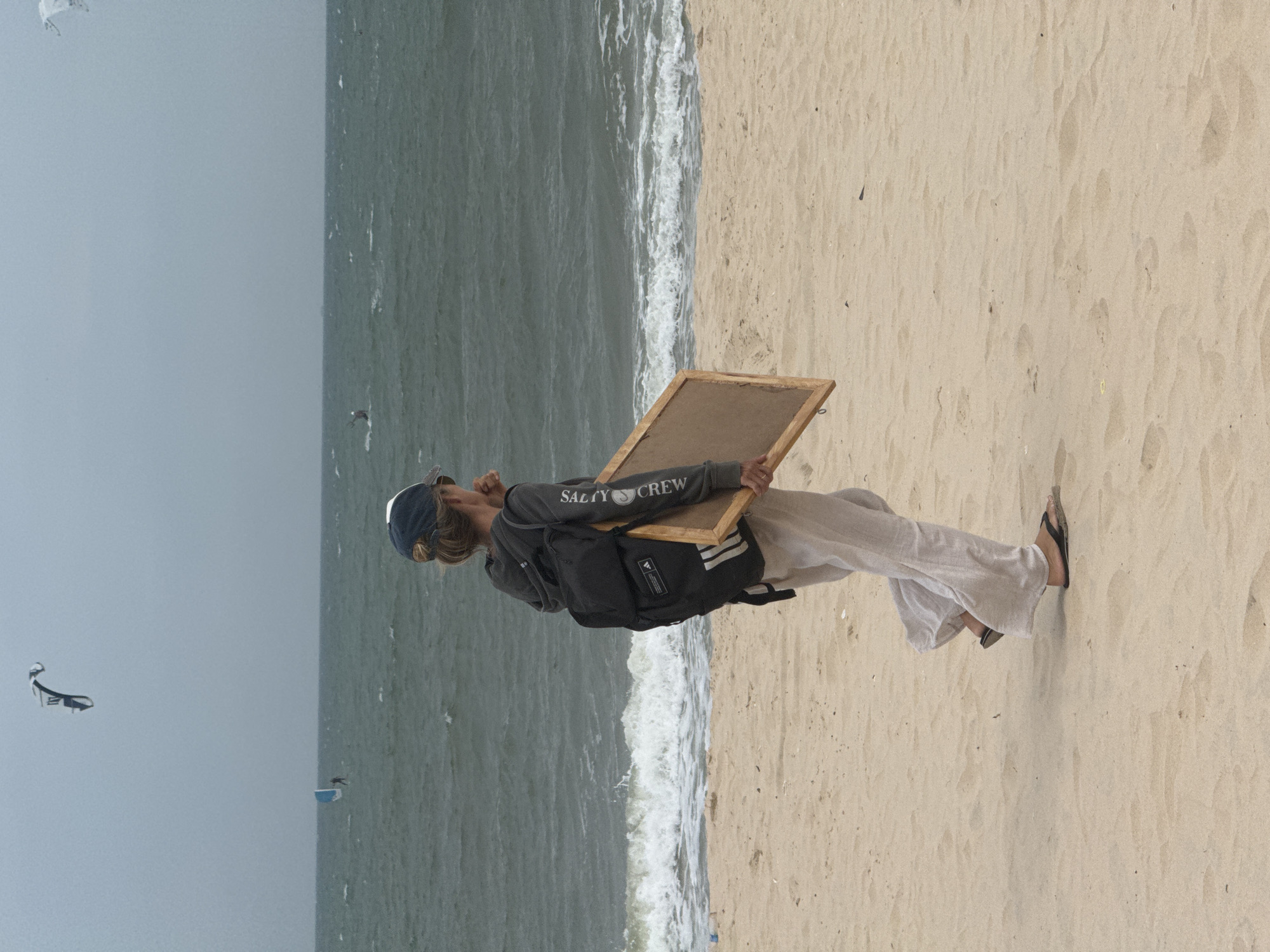A person with a backpack and a wooden frame walks on a sandy beach with the ocean in the background. The sky is overcast. The person is wearing a cap, a grey hooded sweatshirt, and loose-fitting pants. They are carrying a rectangular wooden frame. In the distance, there are kitesurfers on the water. This scene is consistent with the activities and environment of Mũi Né, Vietnam, known for its beaches and watersports.