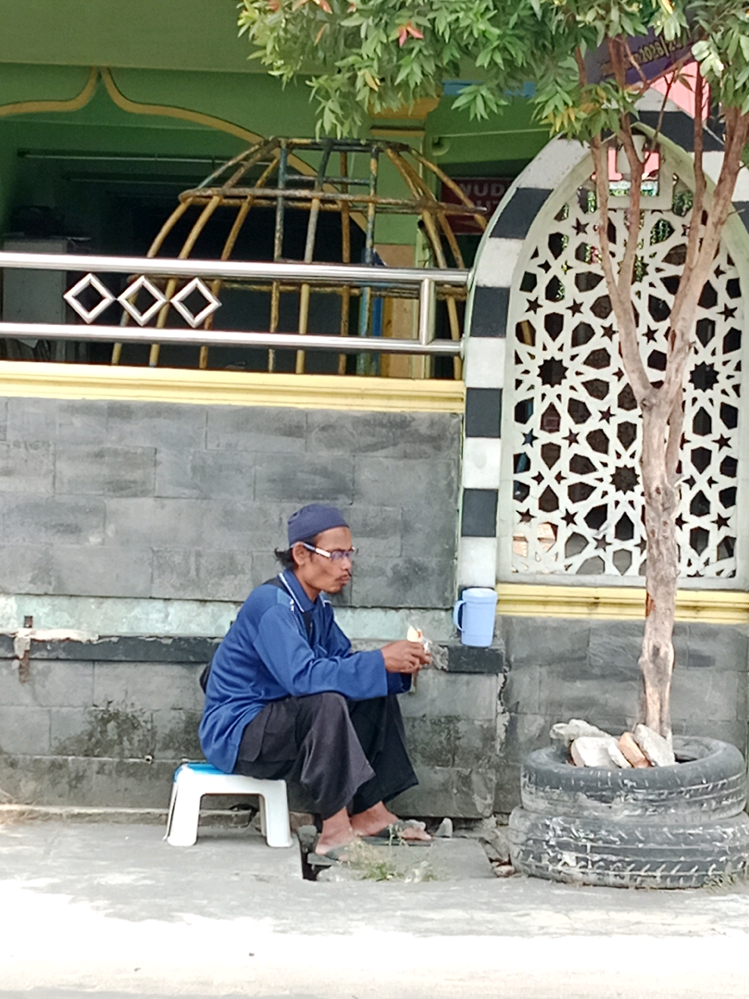 In Cemani, Indonesia, a man wearing a blue long-sleeved shirt, dark pants, a blue cap, and glasses sits on a white stool next to a grey brick wall. He is holding a small white object in his right hand and appears to be eating. To his left, a blue mug sits on a ledge. To his right, a young tree grows out of two stacked tires filled with rocks. Behind the man is a building with a green facade, yellow trim, and a decorative arched window with a black and white patterned frame. A metal railing with diamond shapes runs along the building's facade. Lush green foliage is visible above the building.